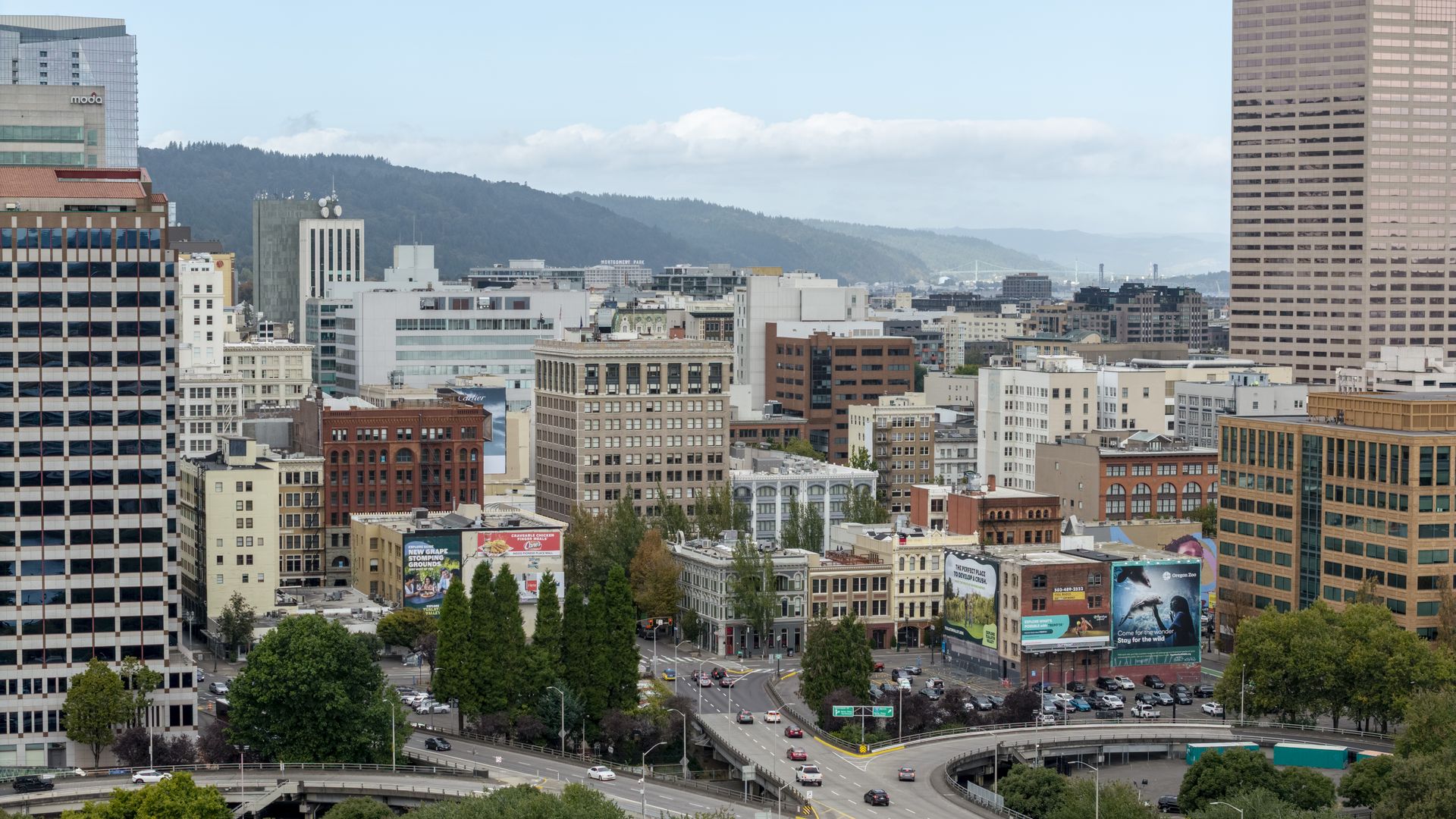 Panoramic cityscape of a dense downtown with glass and brick buildings, a curved highway interchange in the foreground, green trees lining streets, and distant hills beneath a blue sky with clouds.