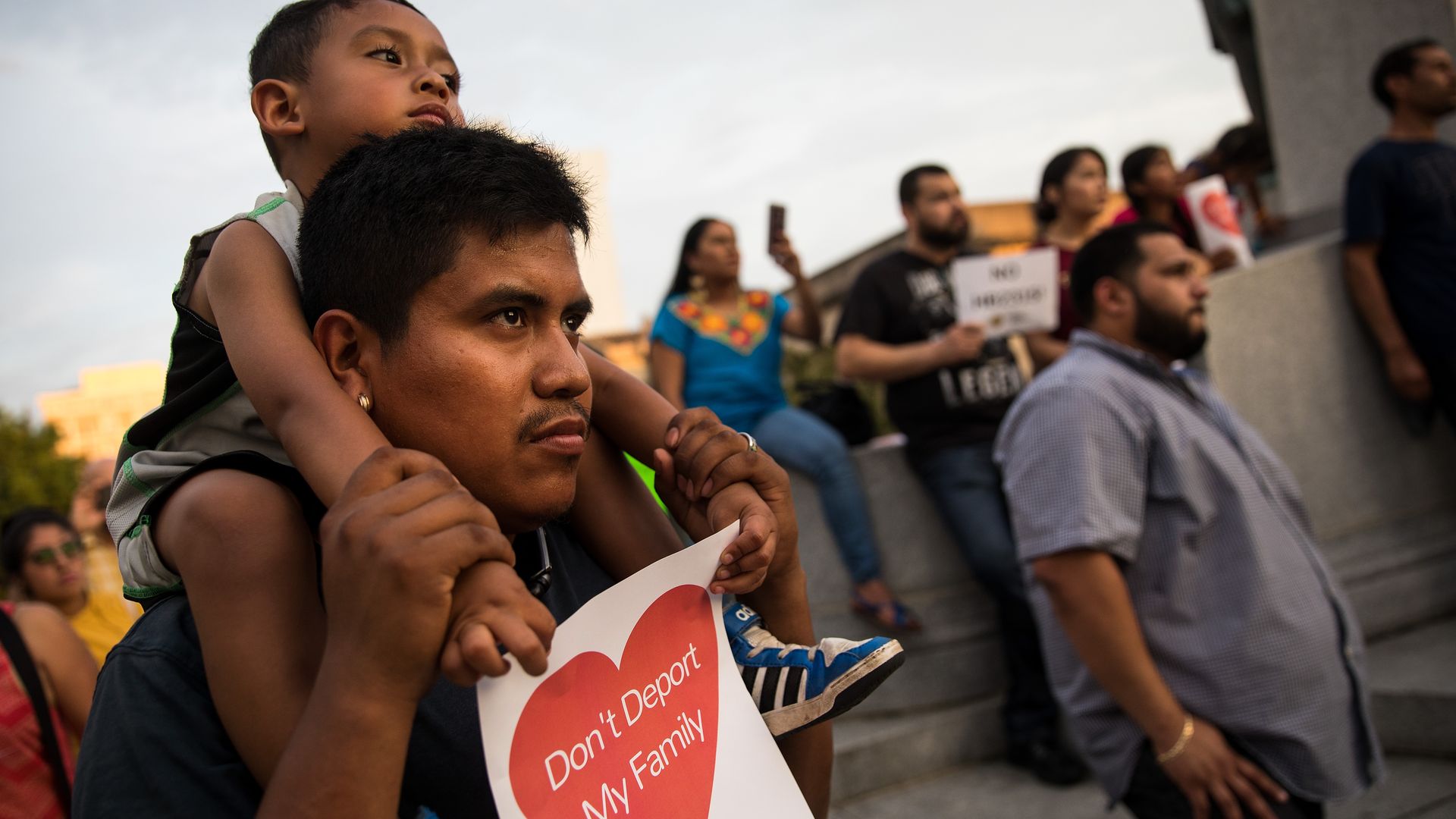 Photo of a young child sitting on an adult's shoulders holding a sign that says "Don't deport my family"