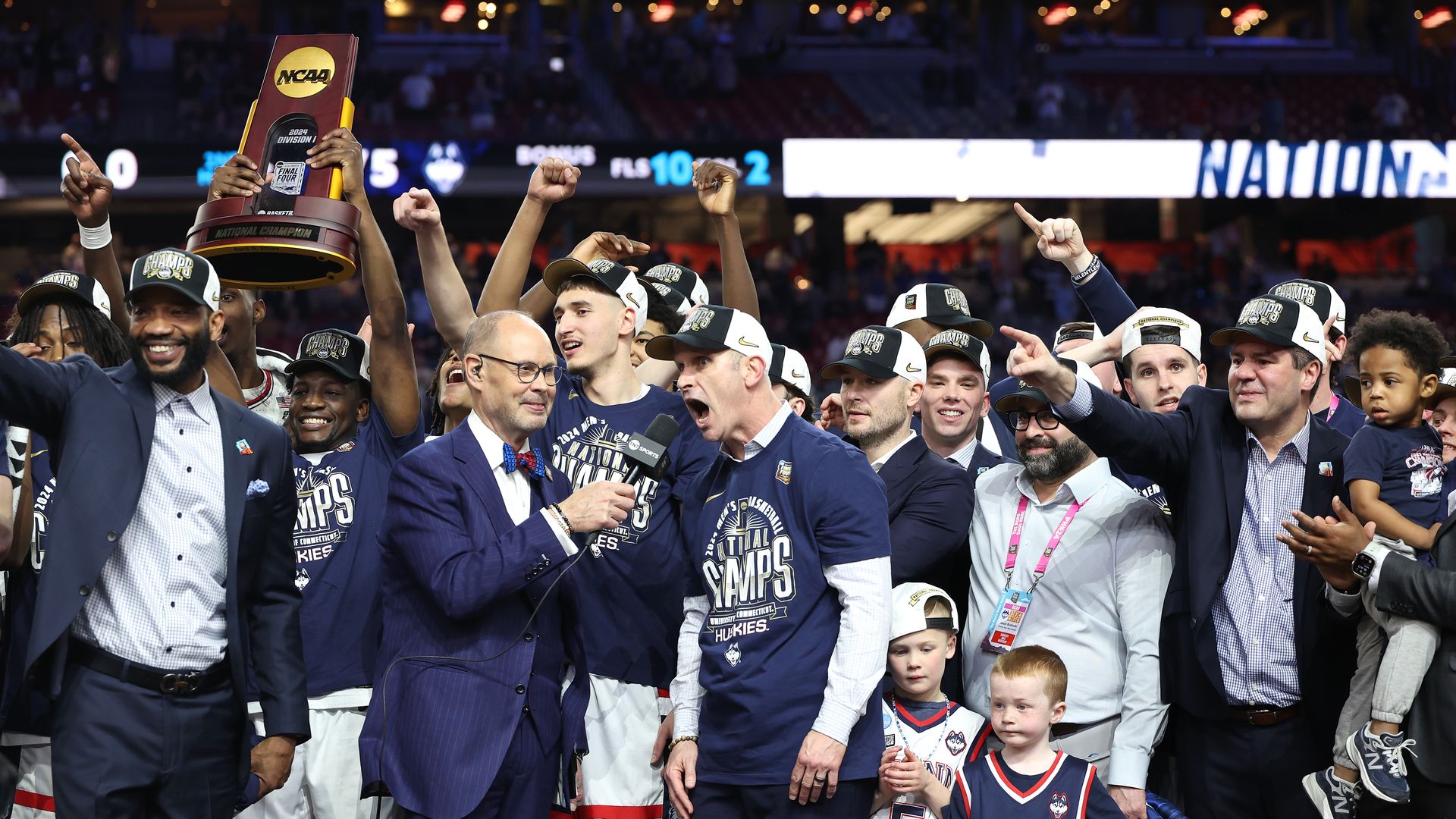 A group of basketball players celebrate on the court and hold up a trophy.