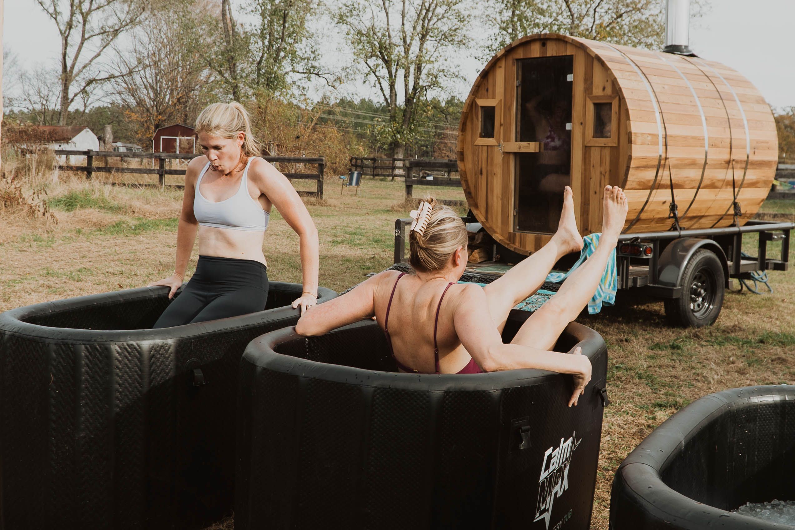Two people in two separate cold plunges facing the opposite way from each other slowly getting in with a sauna in front of them.