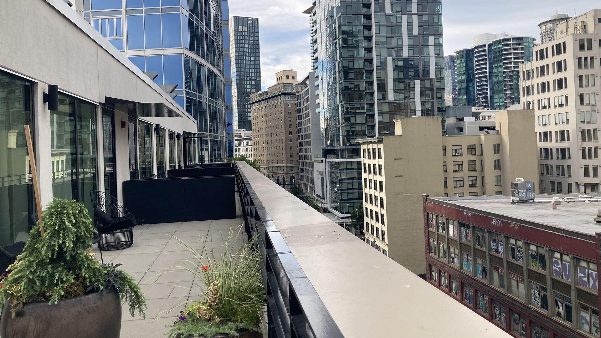 A photo showing a high-rise balcony in a modern city, lined with planters and seating, overlooking tall glass buildings and an older structure.   
