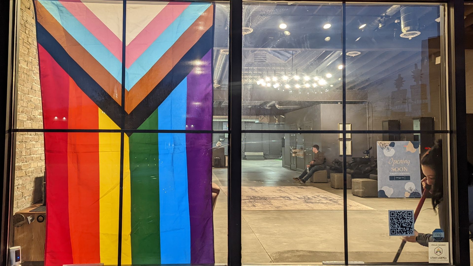 A woman sweeps a salon floor with a Progress Pride flag in the window.