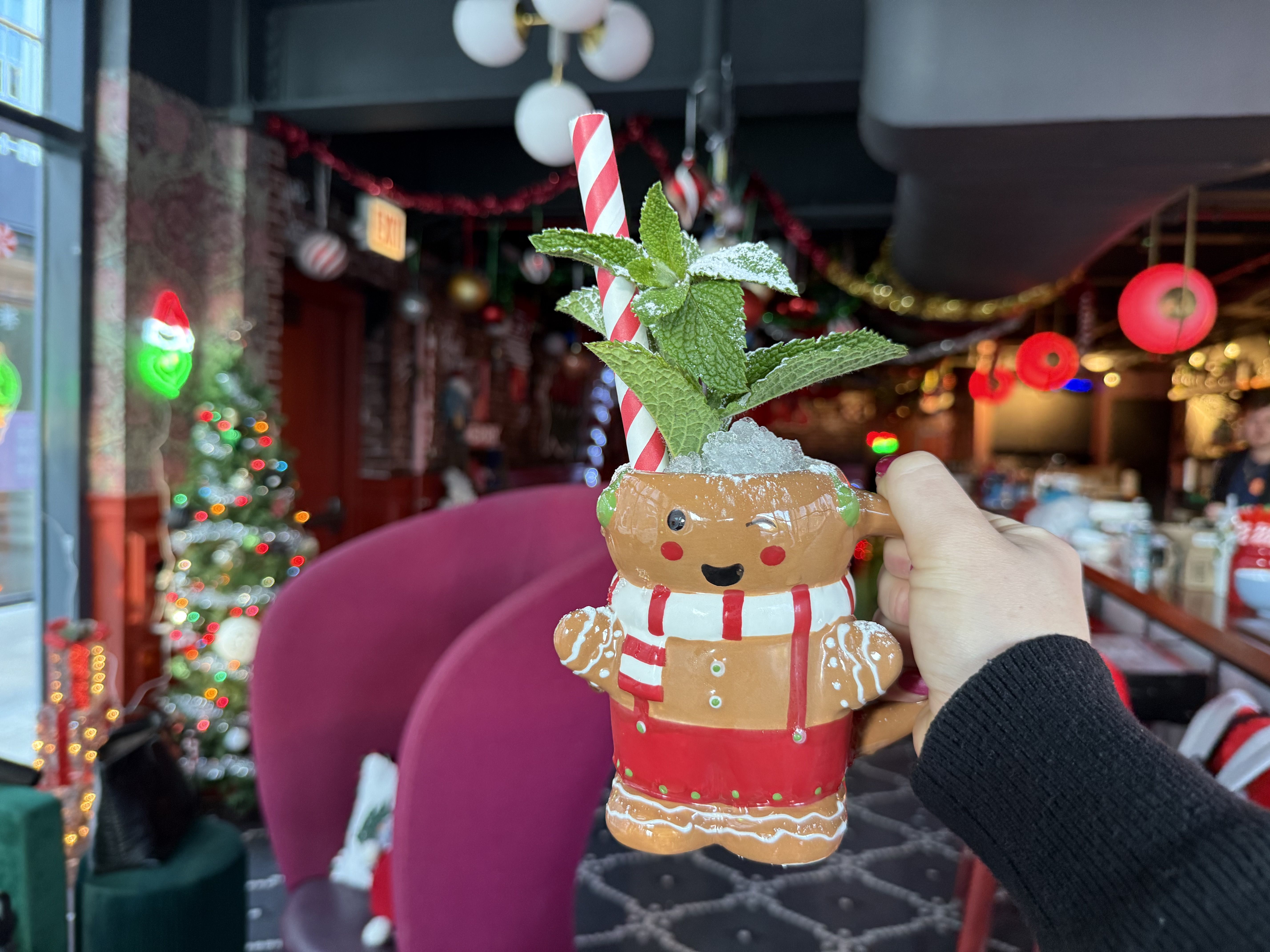 A hand holds a festive gingerbread man mug filled with a drink, crushed ice, red and white striped straw, and mint leaves, in a Christmas-decorated indoor setting with lights and ornaments.