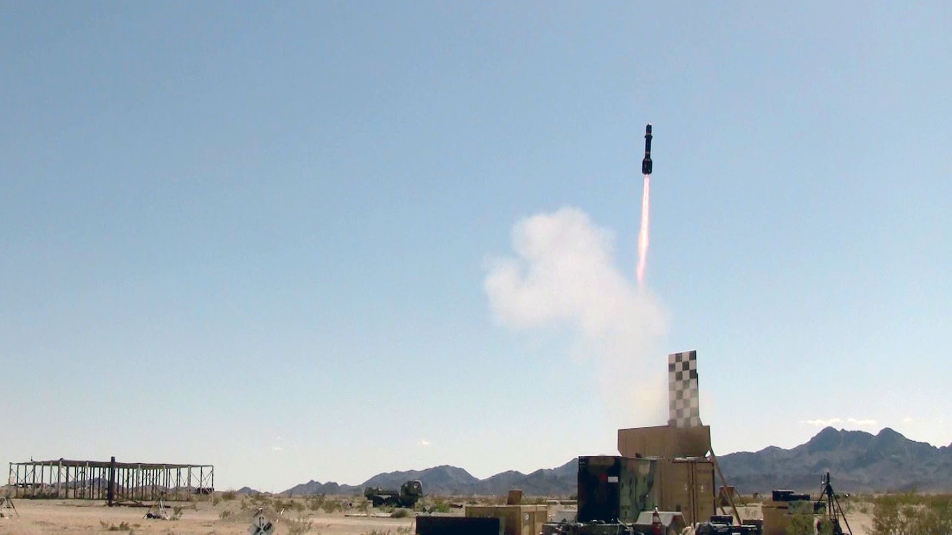 Missile lifts off from a desert test site, sending an orange flame and white plume into a clear blue sky.