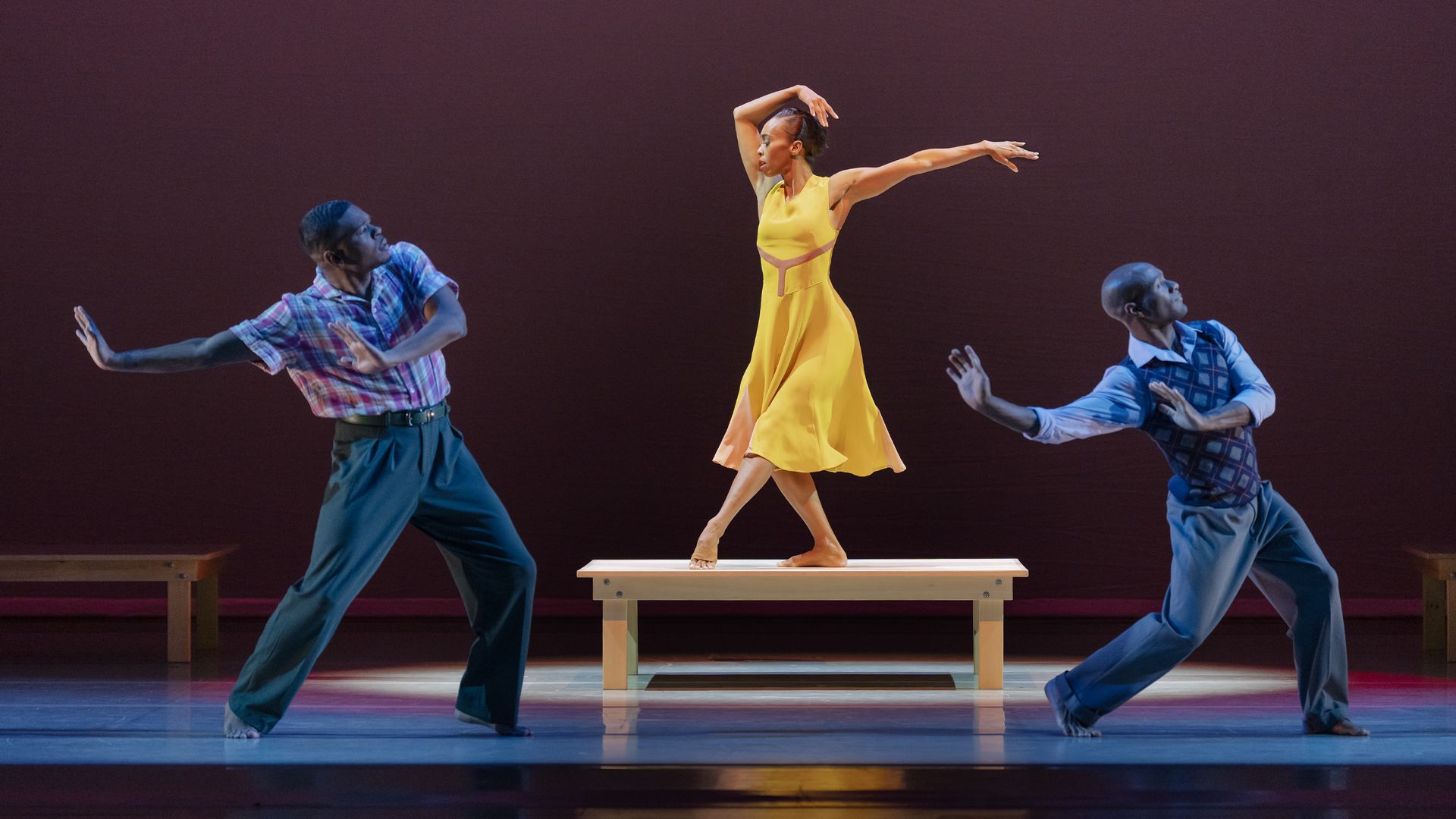 Three dancers on stage with a reddish background, a woman in a yellow dress standing on a wooden bench, flanked by two men in blue pants and patterned tops striking poses.