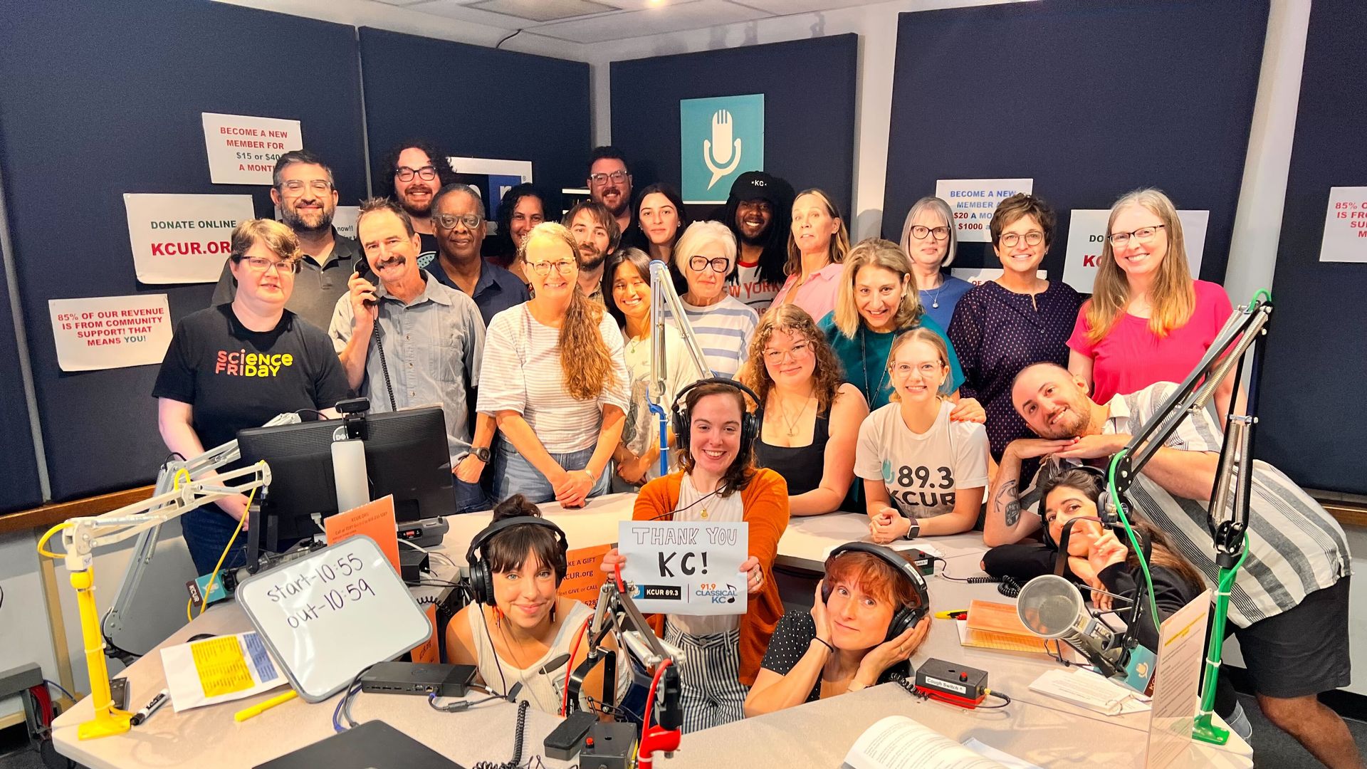 Group of smiling people in a radio studio with microphones and computers, one holds a sign reading "Thank you KC!" Posters on the walls mention KCUR.org and community support. KCUR staffers in the Up To Date studio at the conclusion of our emergency drive.