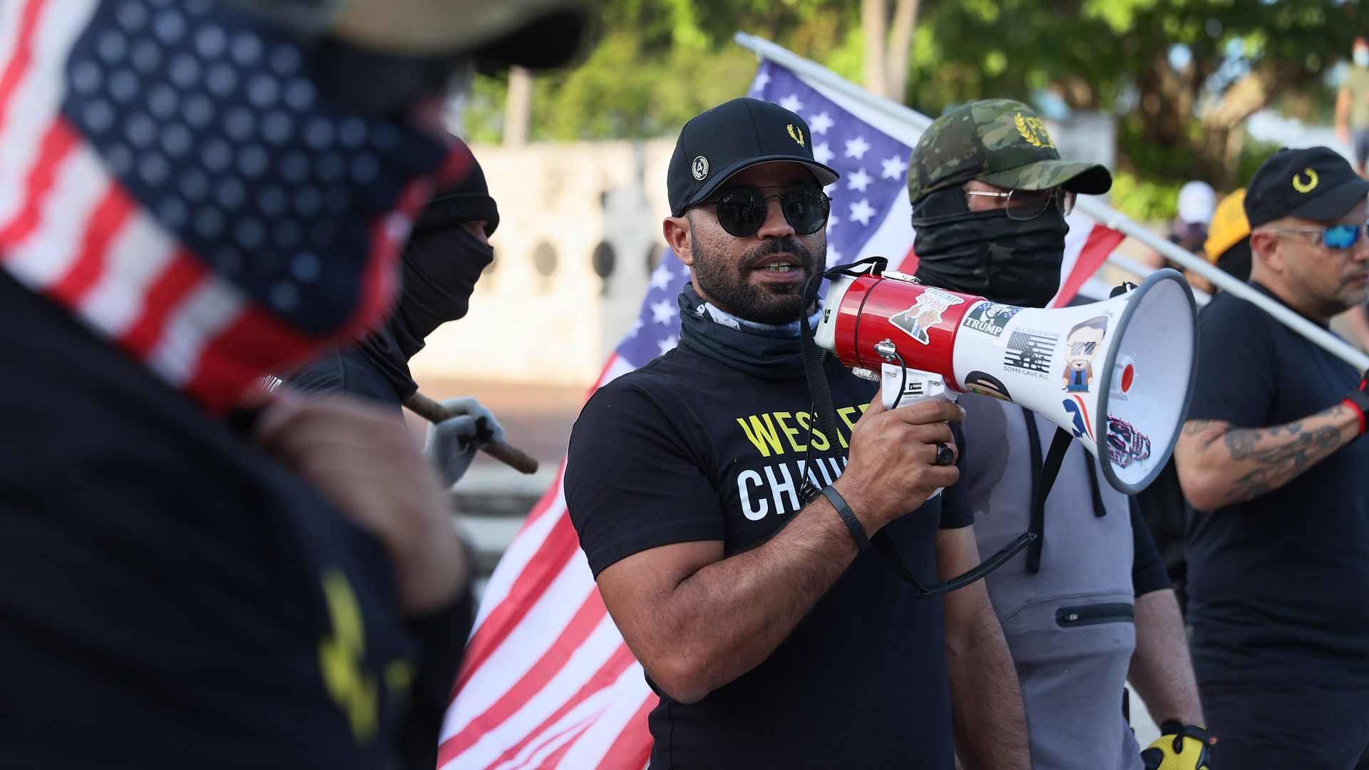 Photo of Enrique Tarrio holding a megaphone and standing with other people with American flags