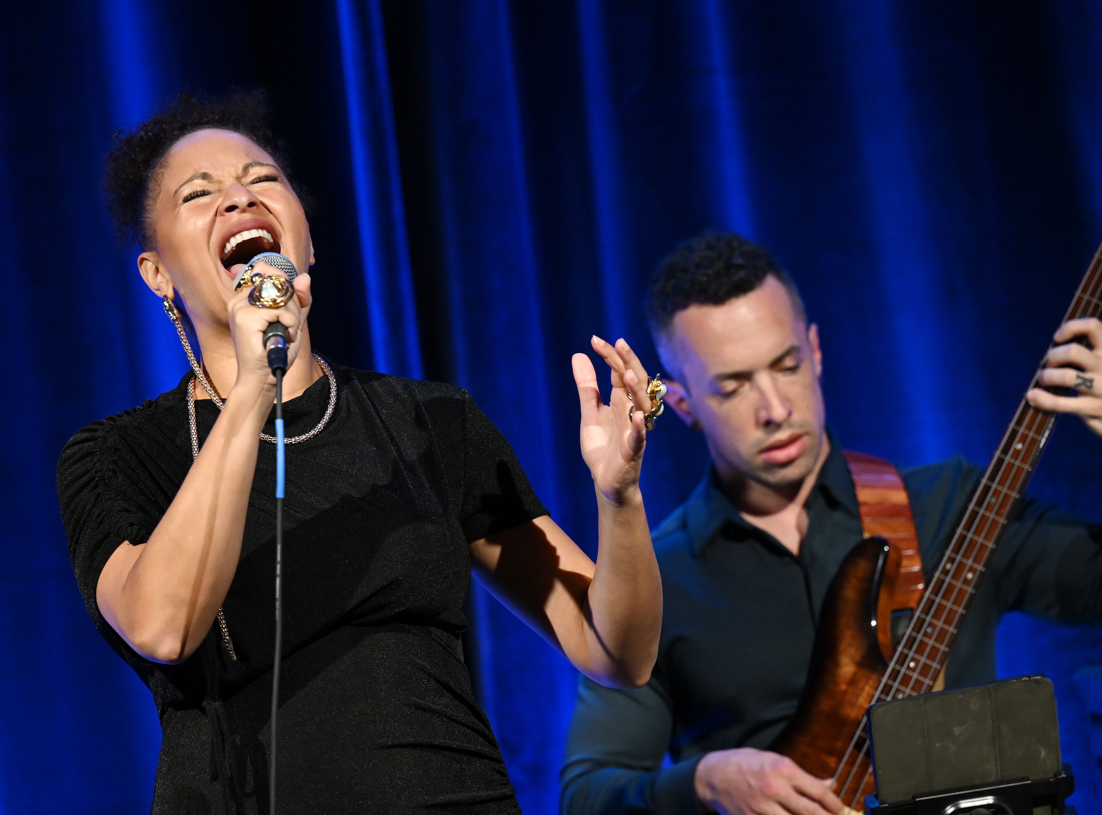 A woman sings into a microphone while a bass player performs behind her.