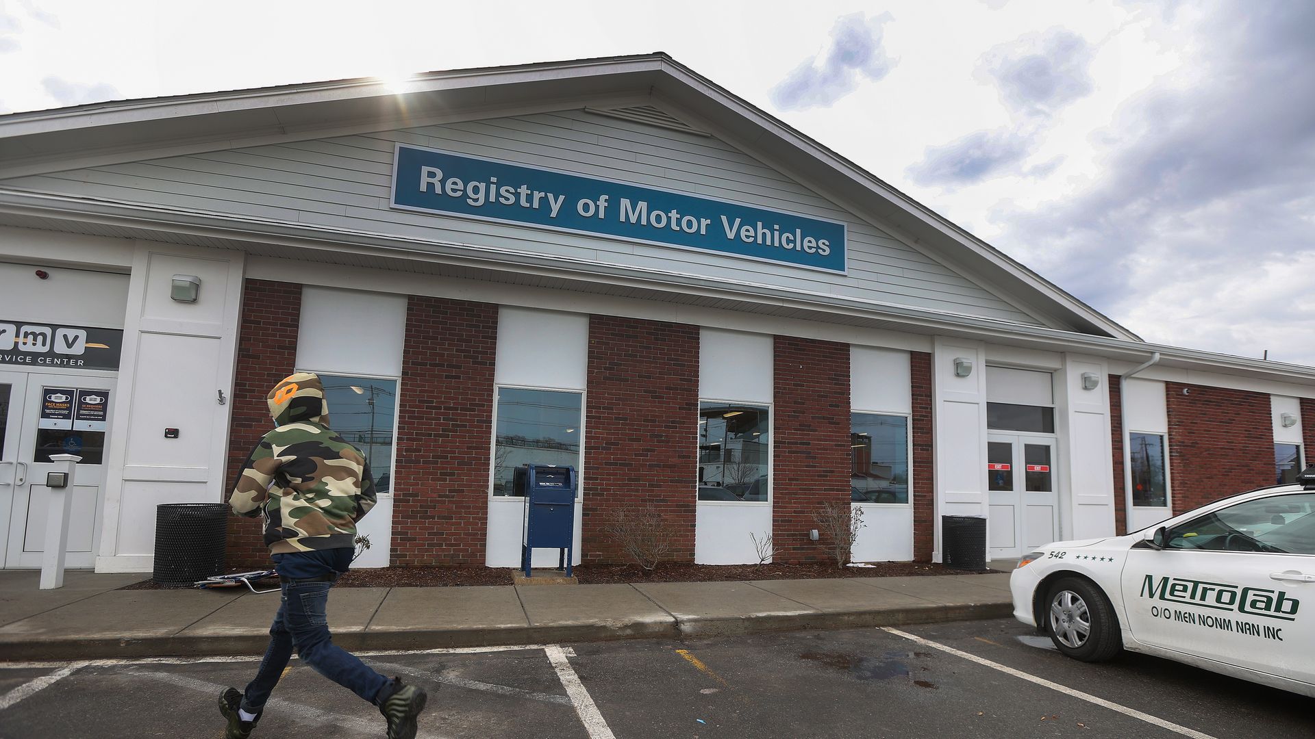A person in an army camouflage jacket walks across the parking lot of a Registry of Motor Vehicles office.