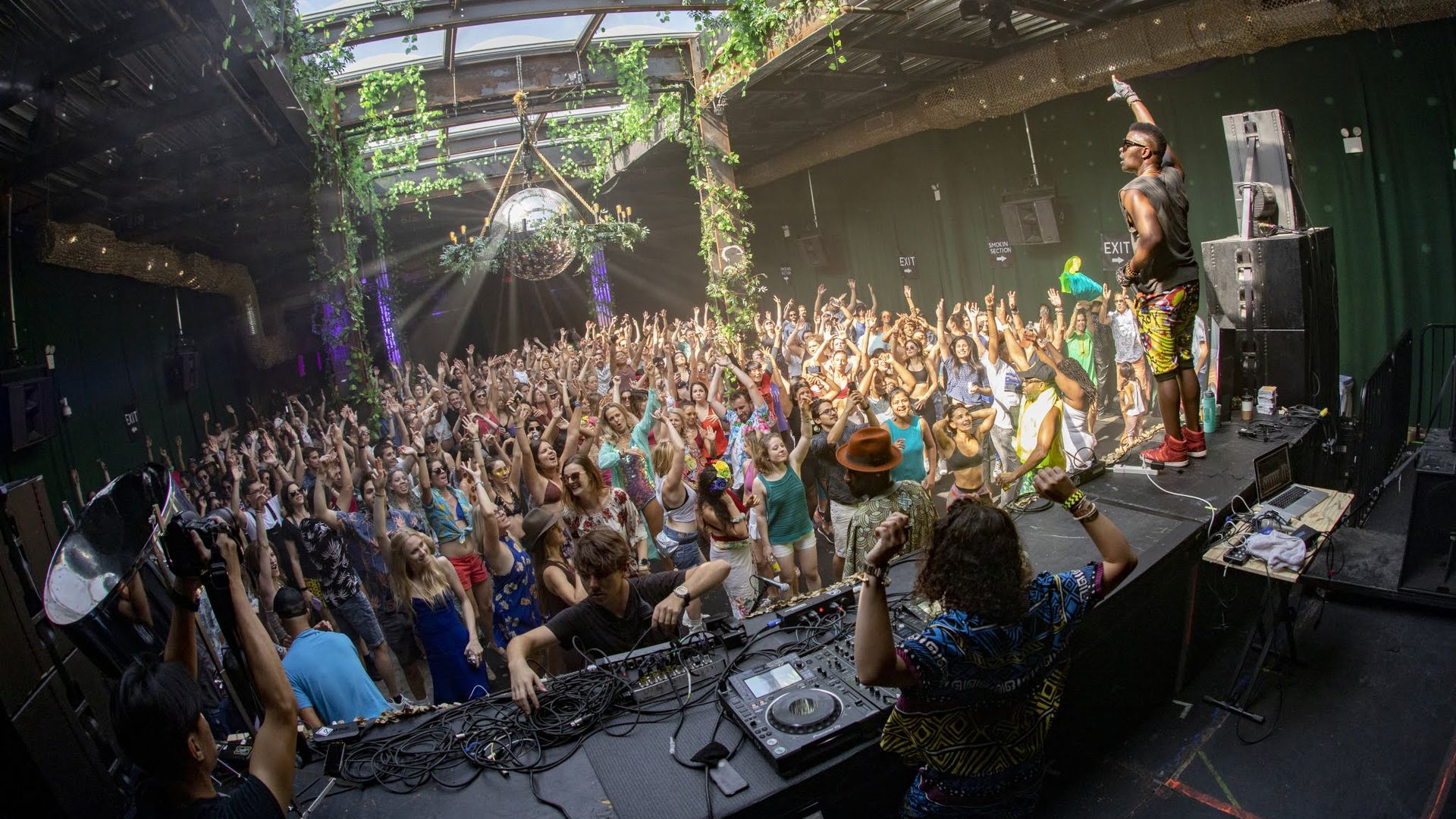DJ and performer energize a large crowd dancing and cheering under a glass roof decorated with hanging green vines and a disco ball.