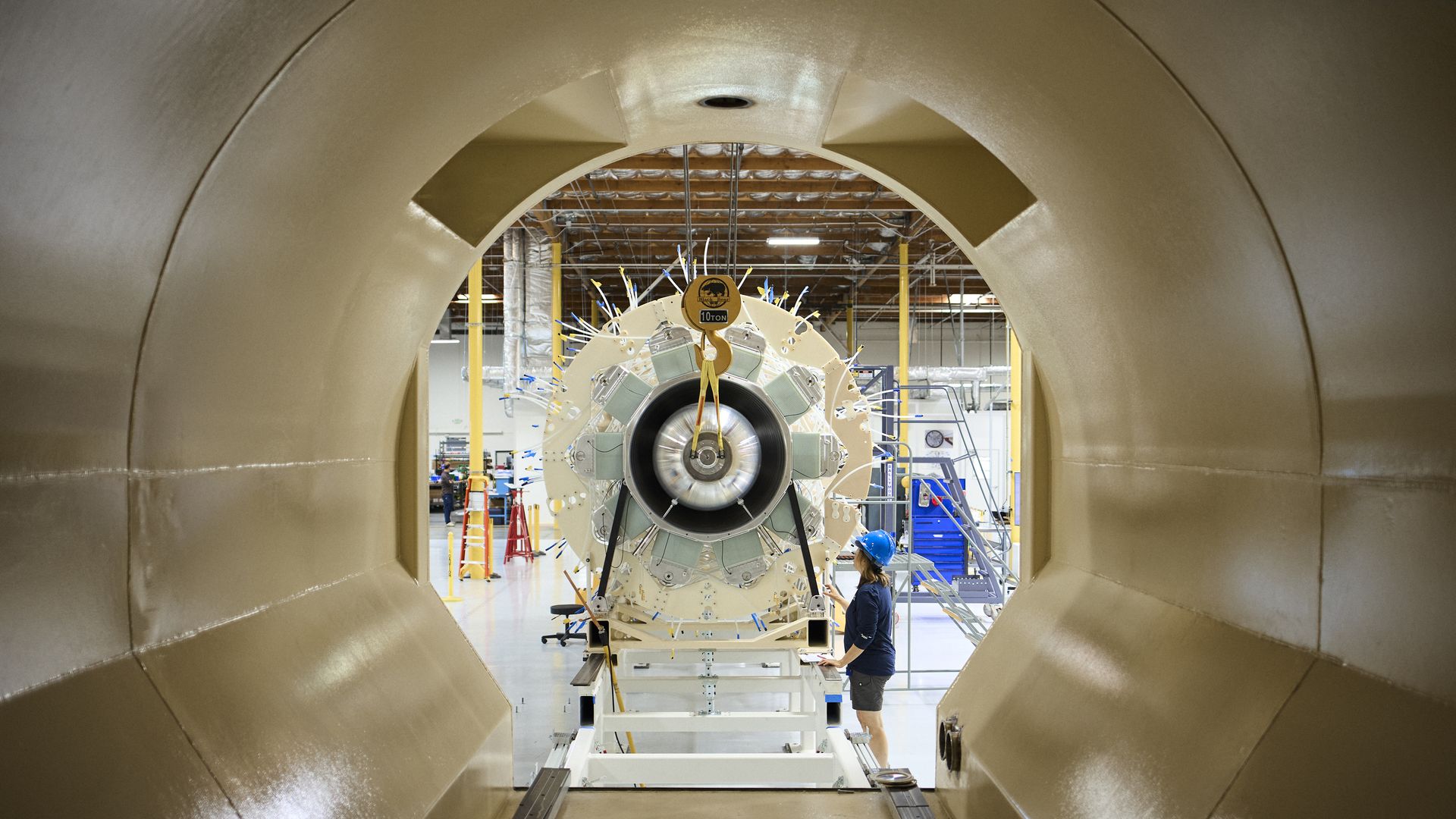 Looking through a beige aircraft fuselage at a jet engine nacelle under assembly; a worker in a blue hard hat stands near yellow supports and cables, with a "10 TON" sign overhead.