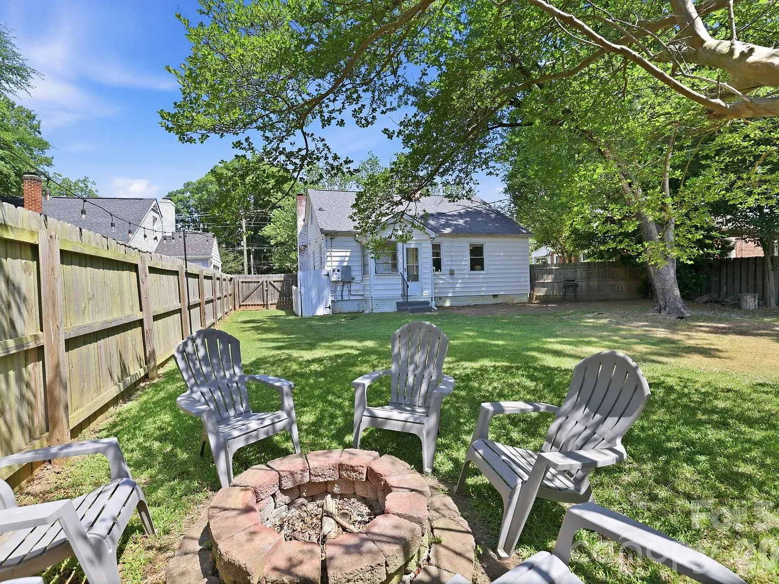 Backyard with a circular brick fire pit surrounded by gray plastic chairs, green grass, a wooden fence with string lights, and a small white house with a gray roof under a large leafy tree.