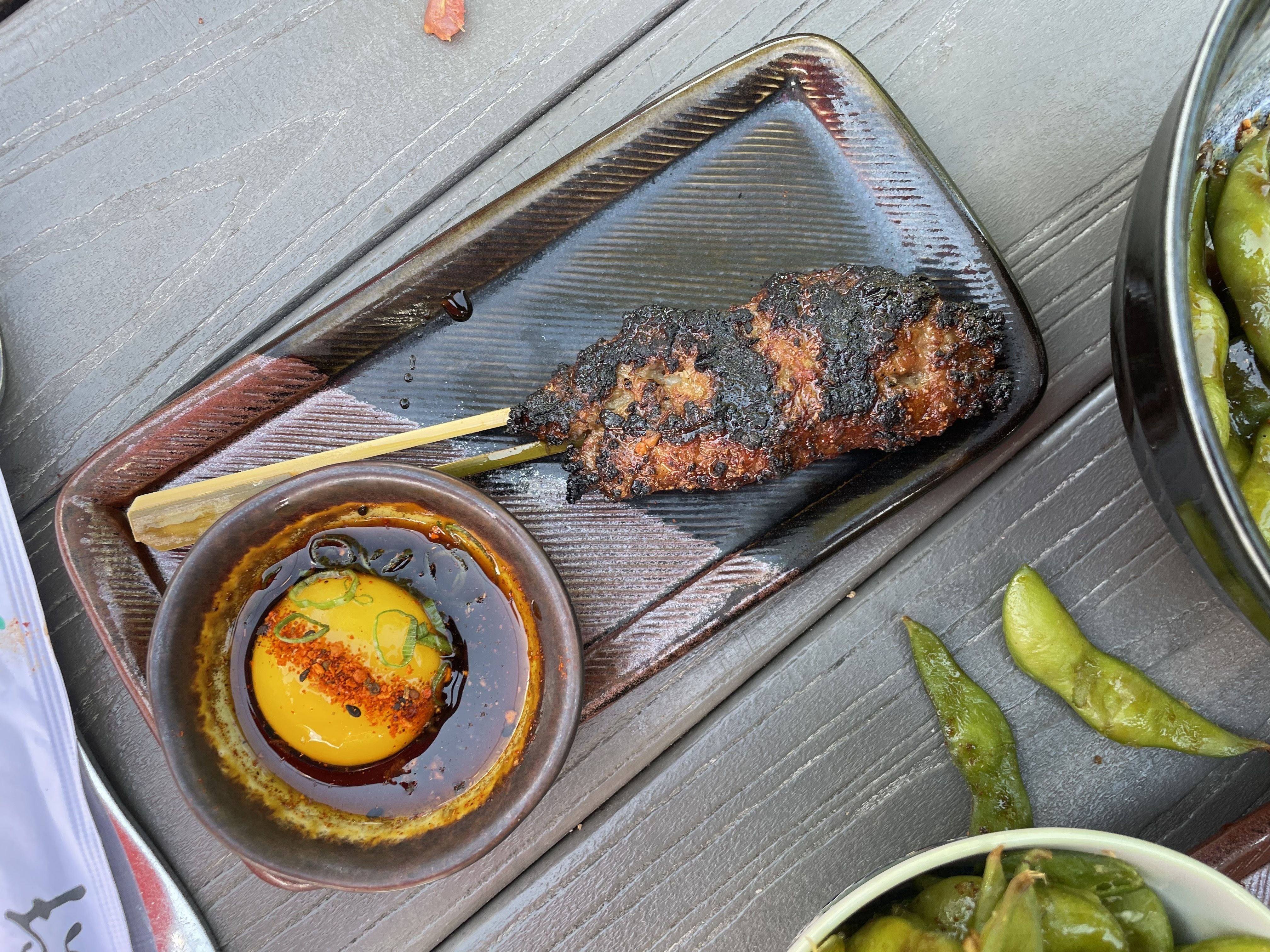 A piece of cooked chicken on skewer on a brown plate next to a small bowl with an egg yolk.