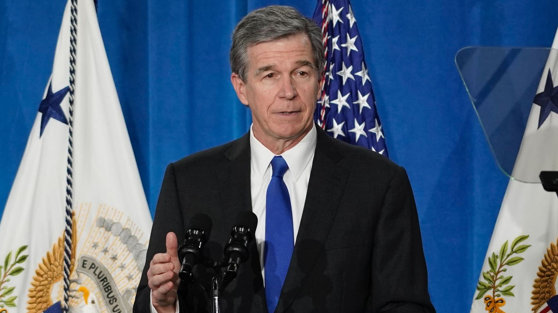 Former N.C. Gov. Roy Cooper, dressed in a black suit with a blue tie, speaking at a podium with the Vice President of the United States seal, with U.S. flags and blue curtains behind him.
