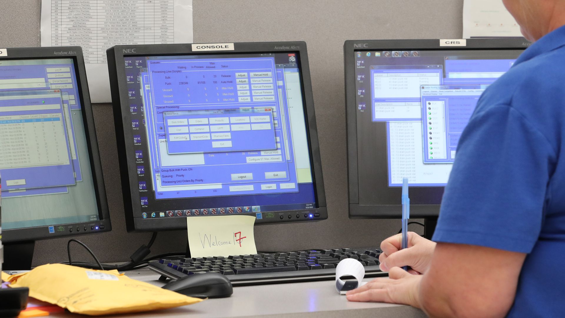 A pharmacy technician writes on a piece of paper in front of three computer screens.