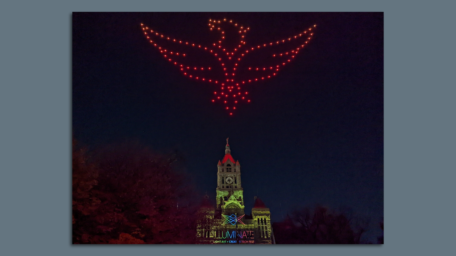 A light-up bird appears in the lights of drones above a bell tower on a tall building.
