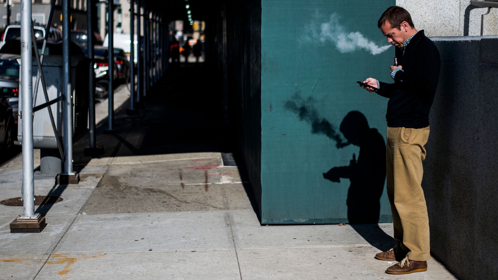 In this image, a man stands and looks at his phone while vaping on the sidewalk. 