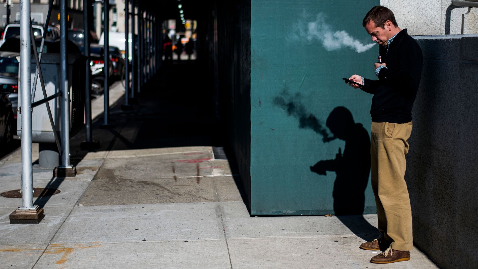In this image, a man stands and looks at his phone while vaping on the sidewalk.