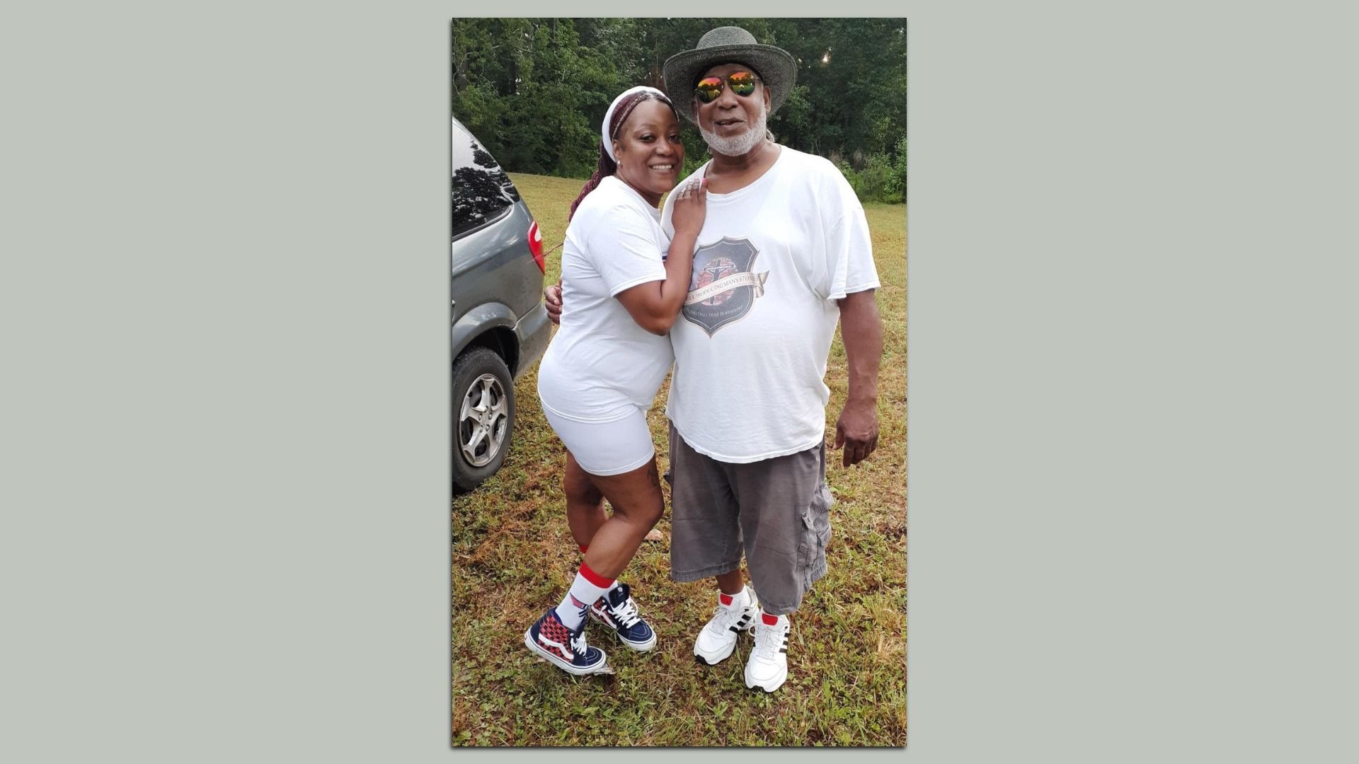 A smiling woman in white shorts and t-shirt with red and blue shoes hugs an older man in a white t-shirt, gray shorts, hat, and sunglasses on grass with trees and a gray vehicle nearby.