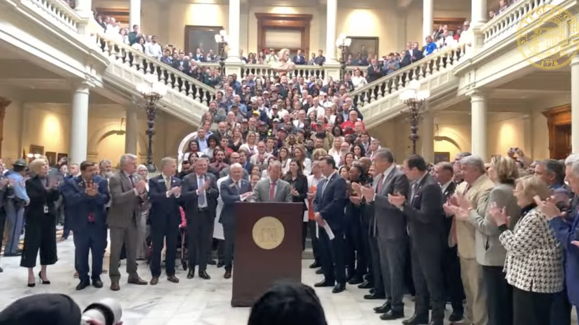 An individual wearing a suit stands at a lectern surrounded by clapping lawmakers standing on a stairwell in a state government building