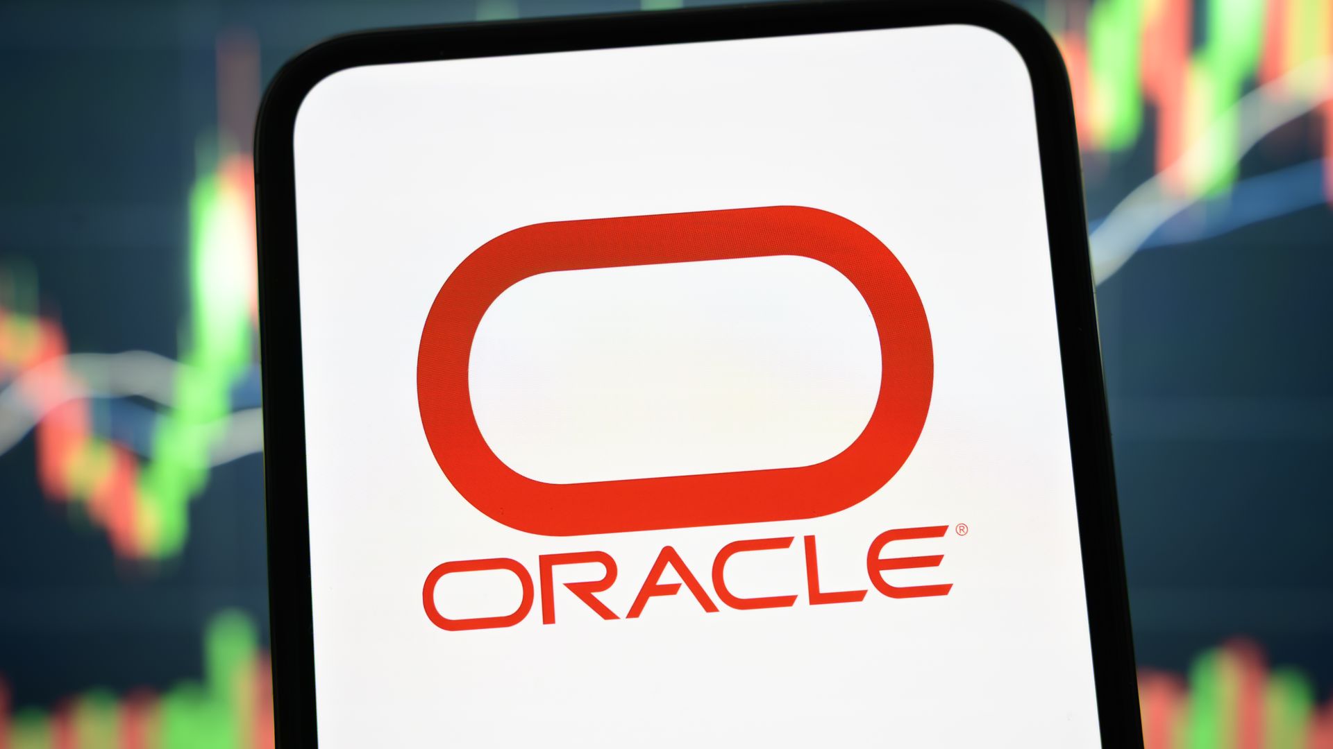 Close-up of a smartphone screen displaying the red Oracle logo on white, with blurred stock charts in the background.