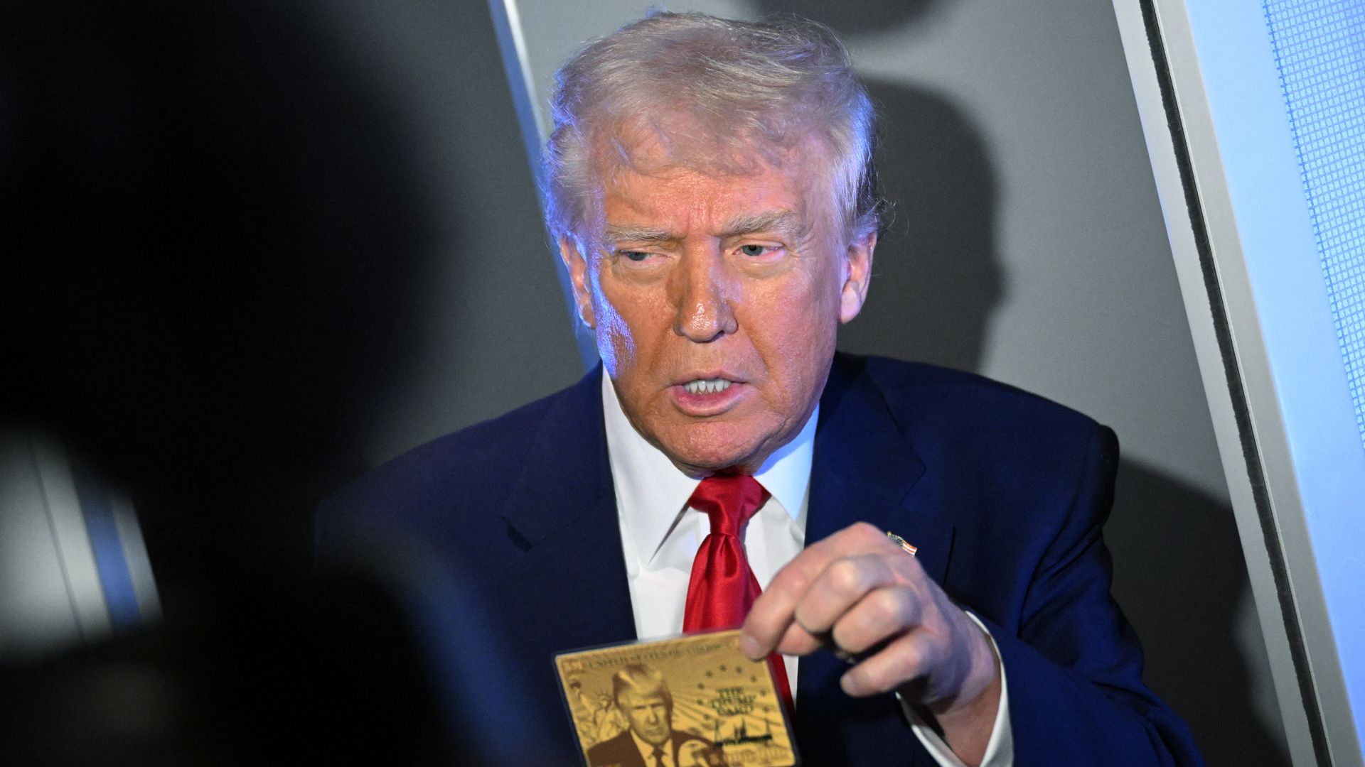 President Trump, wearing a navy jacket with a US flag pin at the top of his left lapel, white shirt and red tie, holds the Trump Gold Card, containing an image of himself in similar dress, while in flight on board Air Force One, en route to Florida .
