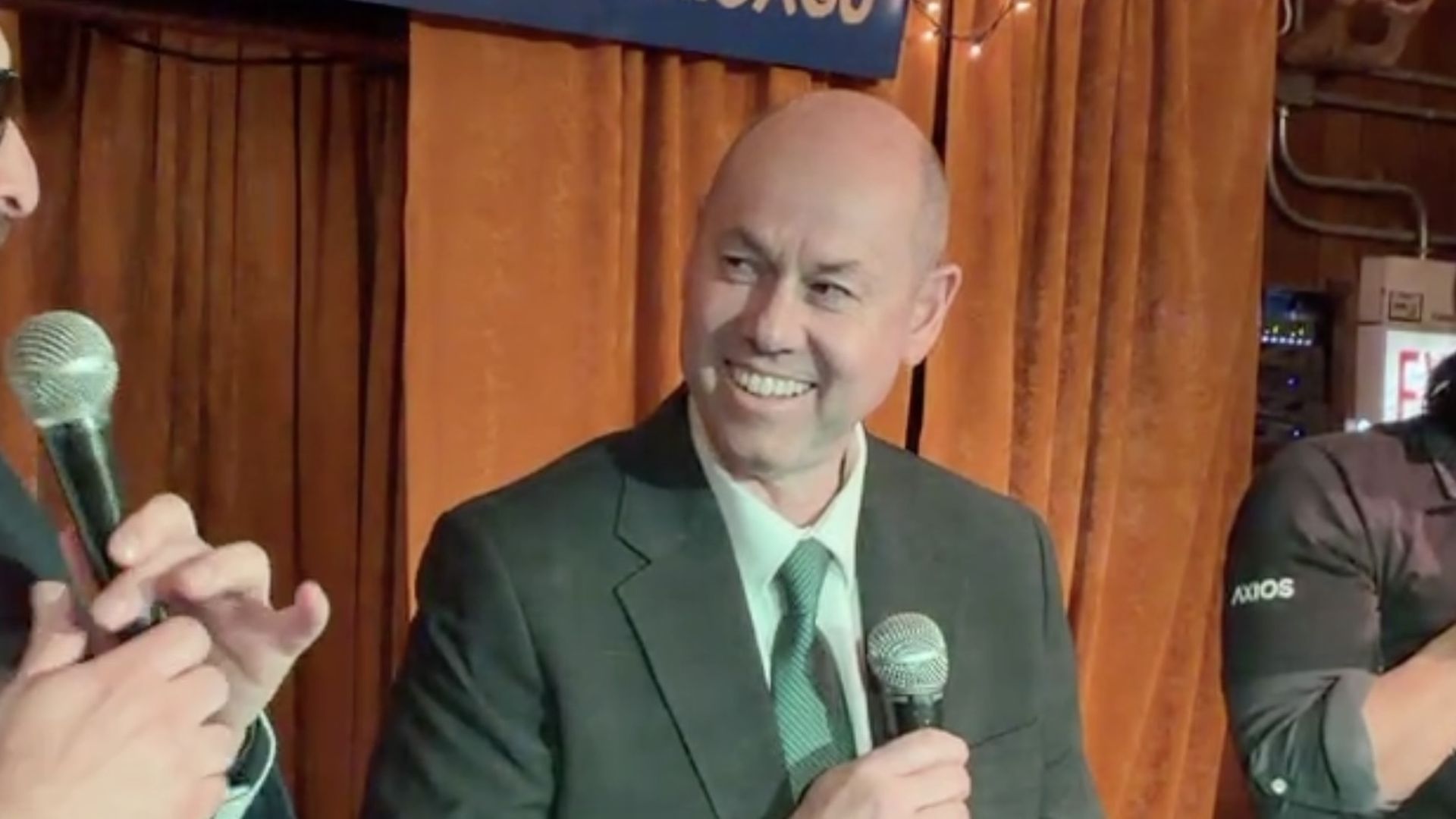 A balding man in a dark suit and green tie smiles while holding a microphone on a stage with orange curtains; another person with a mic is on the left, and a banner above reads "CHICAGO".