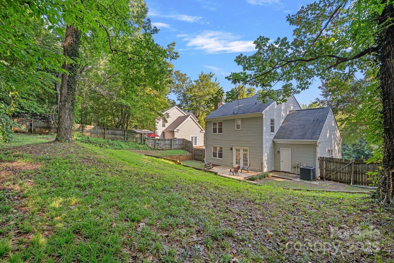Backyard view of a two-story beige house with a small patio holding two chairs, surrounded by green grass, trees, wooden fence, and a blue sky with scattered clouds.
