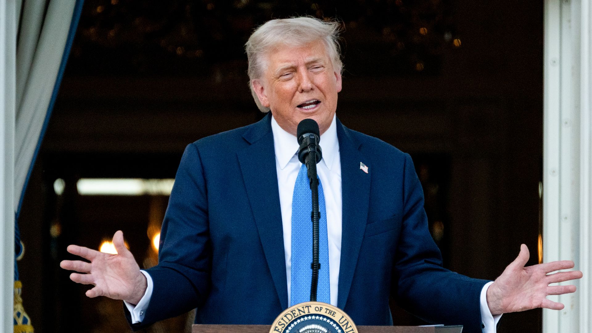 Trump speaks at a lectern with hands outstretched. He is wearing a blue suit, white shirt and blue tie. 