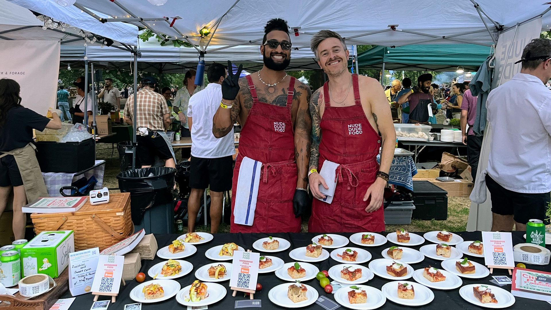 Two men wearing red aprons labeled "Nude Dude Food" stand behind a table with multiple small plates of food under a white canopy at an outdoor event with other attendees in the background.