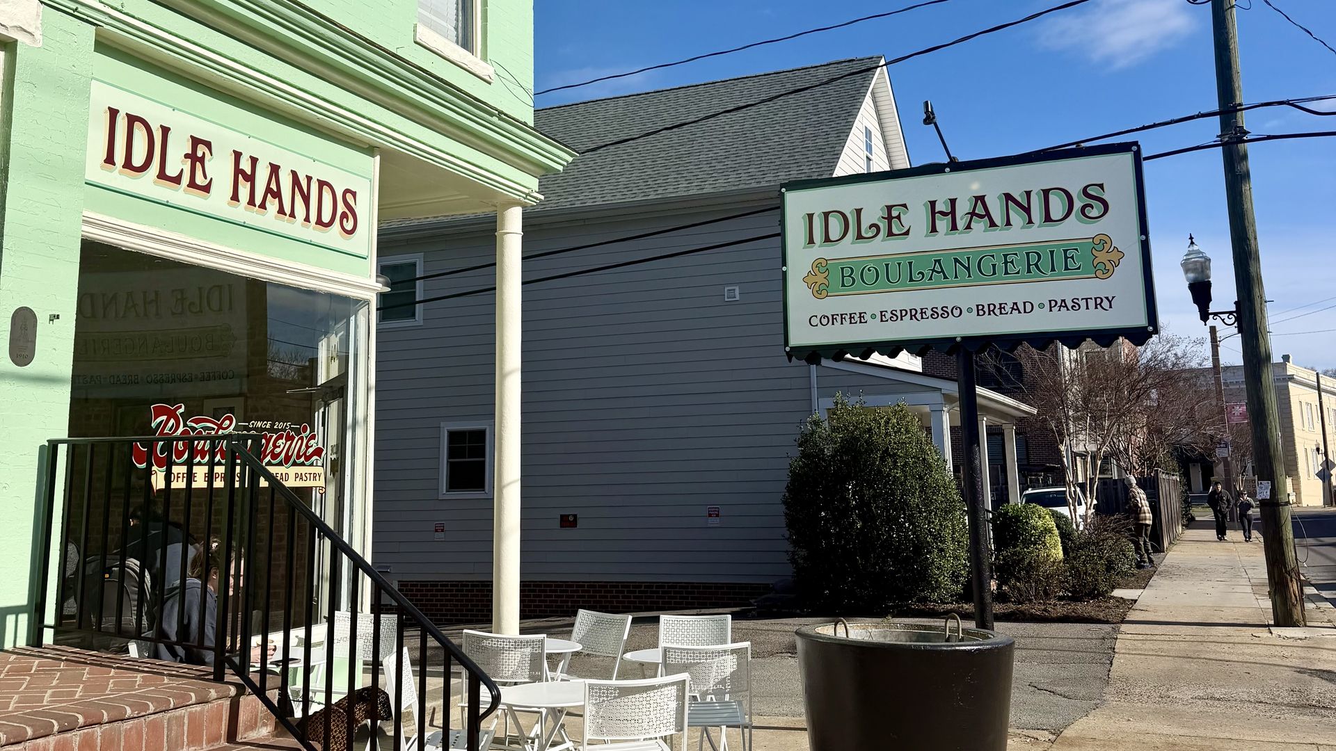 Pastel green, two-story storefront reading "IDLE HANDS" with white outdoor tables and chairs on brick steps; a sign reads "IDLE HANDS BOULANGERIE" under a bright blue sky with power lines.