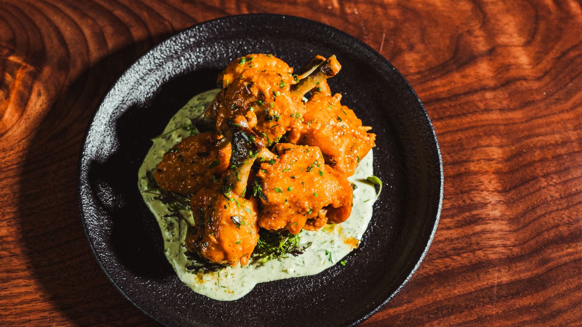 Orange-glazed chicken bites with herbs rest on a pale green herb sauce in a black plate, set on a dark wooden table.