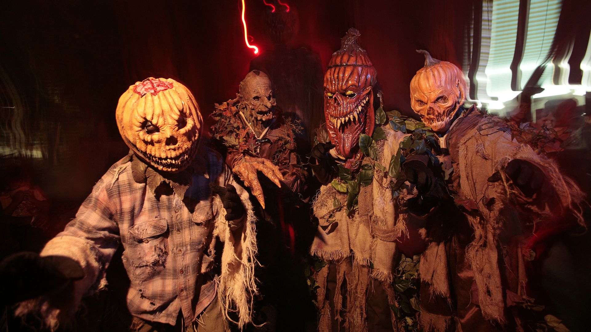 Group of four zombie figures in tattered clothes with pumpkin and skull faces, under dark red lighting, with neon red glow on the left and striped light patterns on the right.