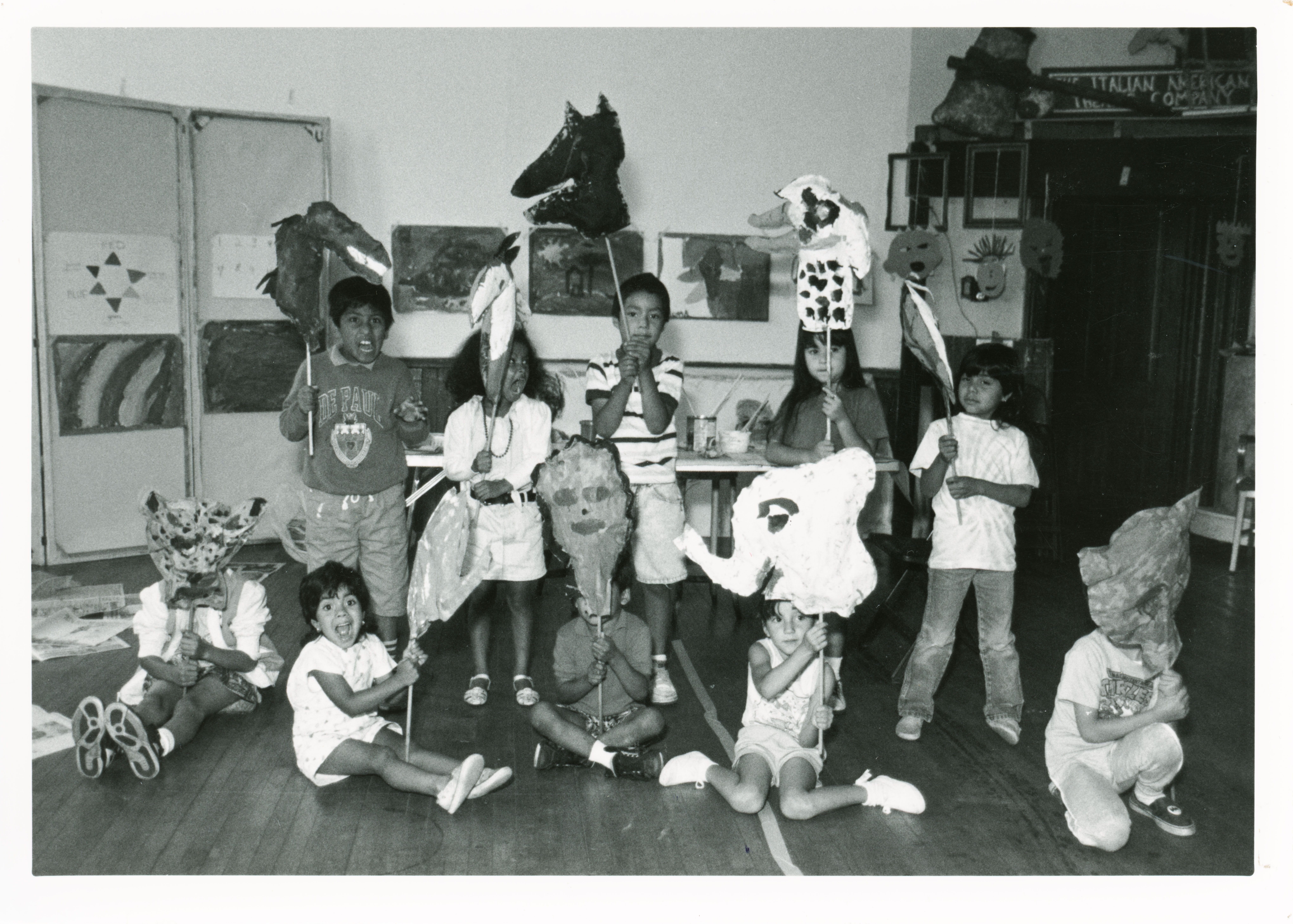 Black and white photo of children in a classroom holding large handmade animal and face masks on sticks, some children sit on the floor while others stand against a wall with artwork.