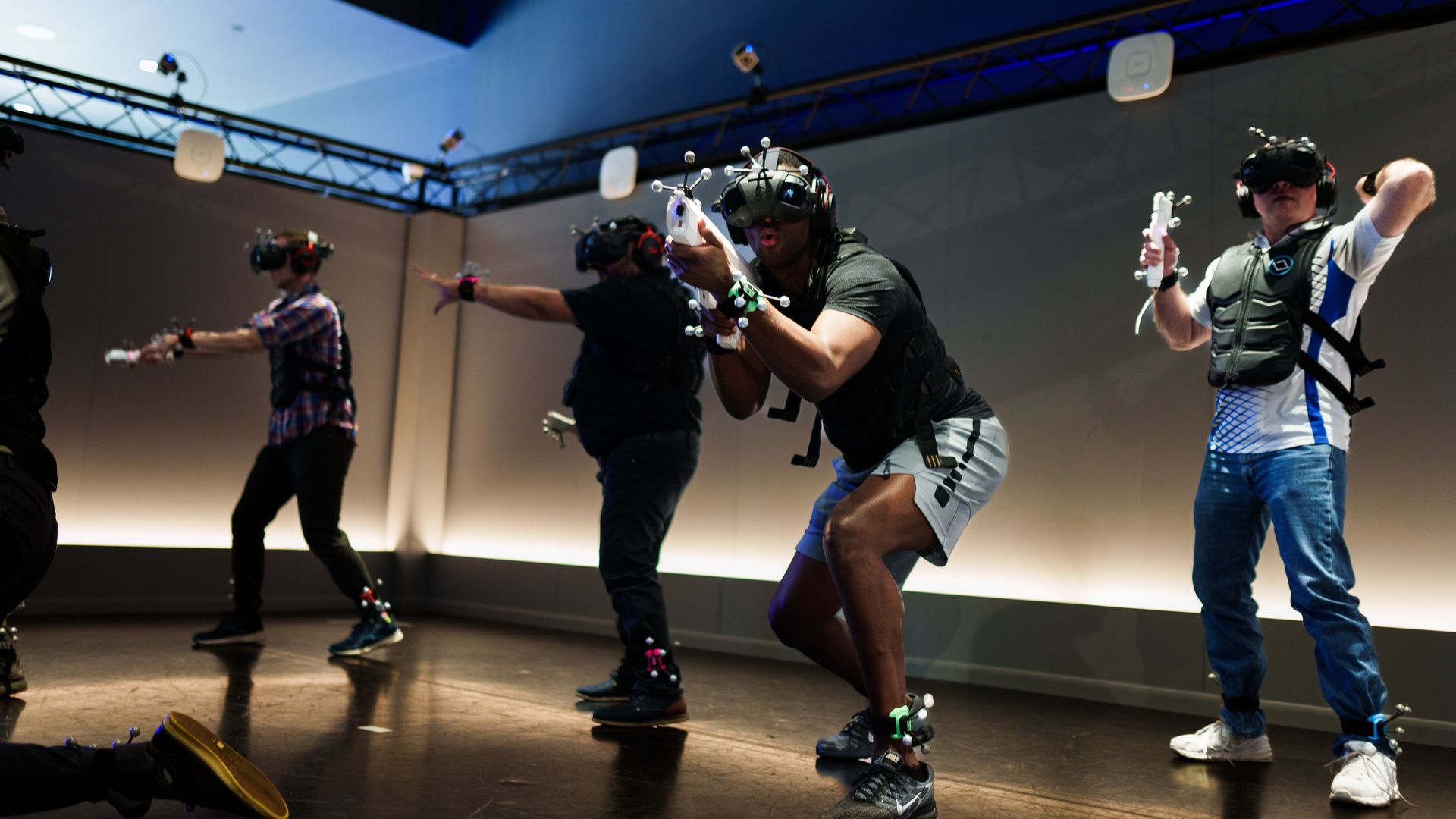 Four people wearing VR headsets, motion capture markers, and vests are engaged in an immersive VR game in a dimly lit room with blue and neutral lighting.