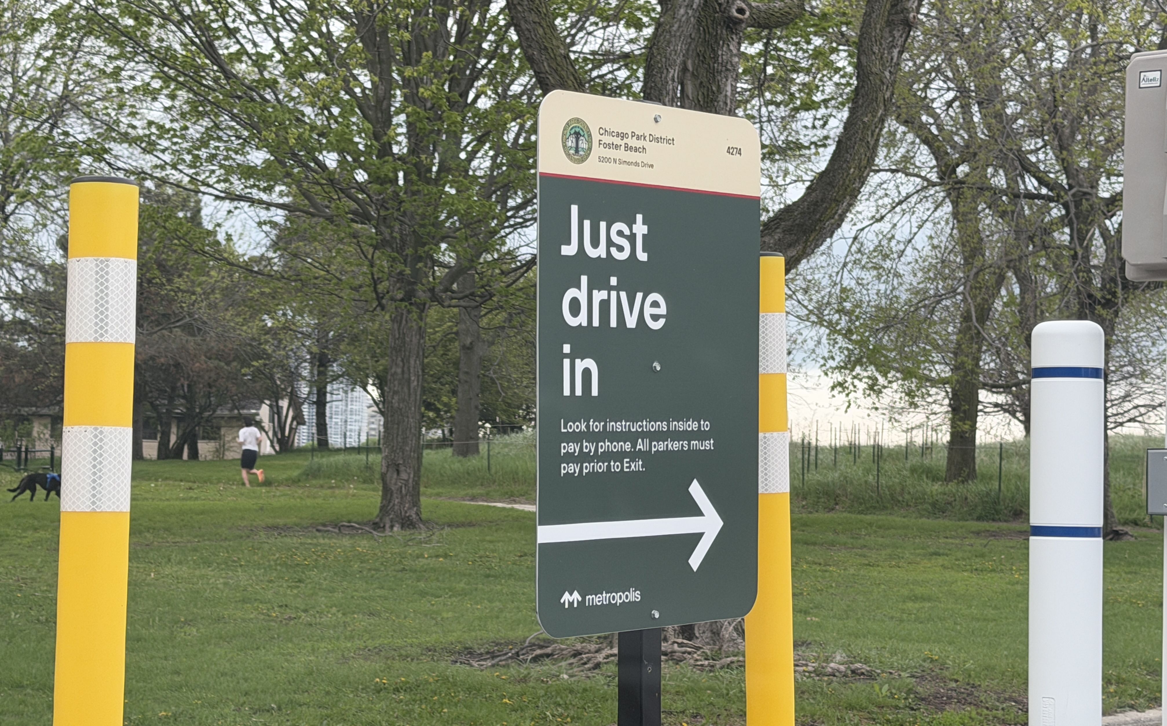 A large sign reads "Just drive in" with an arrow, set in a park. Background shows trees, grass, yellow striped bollards, and a person jogging with a dog.