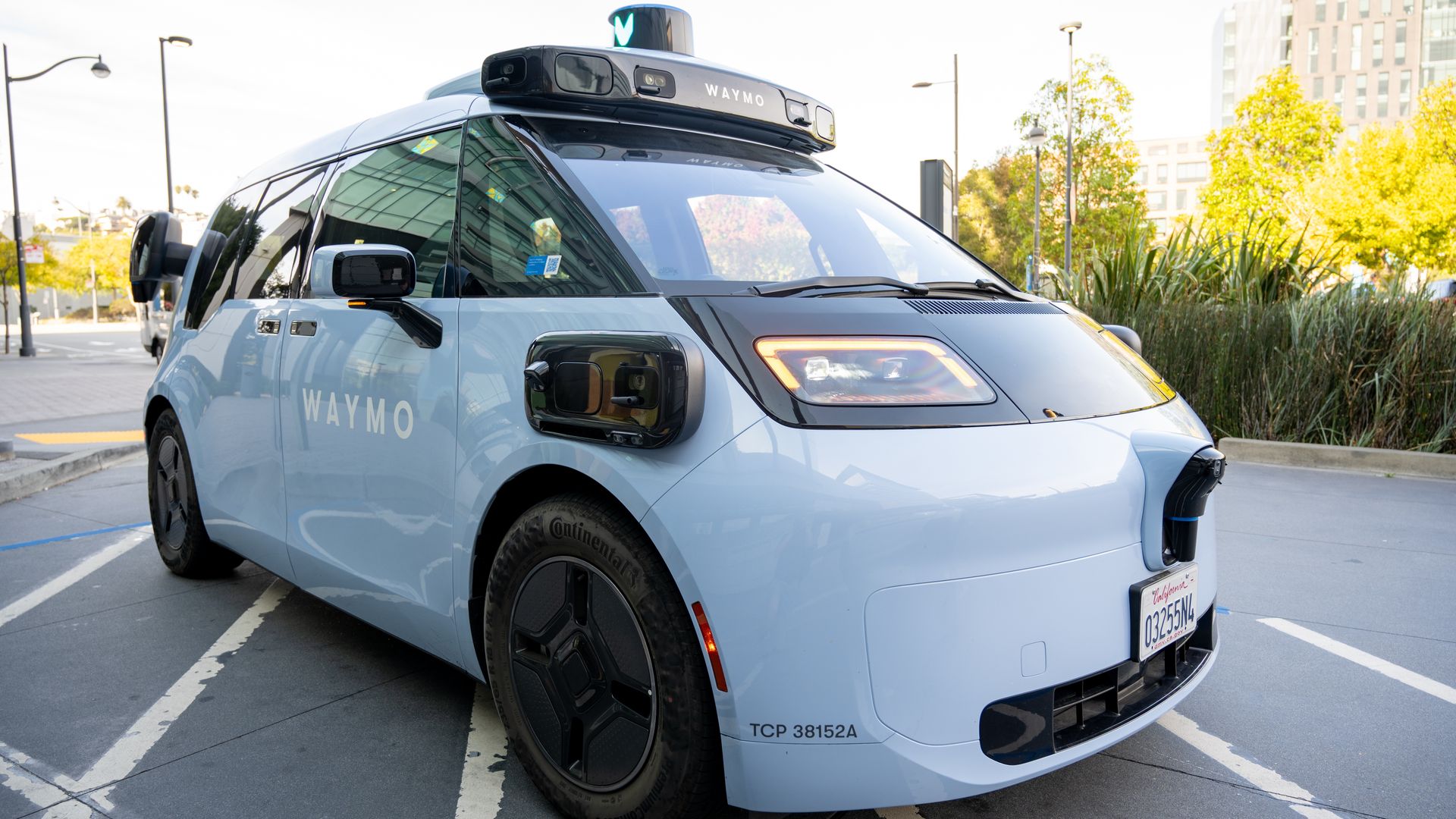 Light blue Waymo autonomous van with roof lidar module and green beacon, parked in a sunny urban lot. WAYMO visible on roof, California plates, modern buildings and greenery in background.