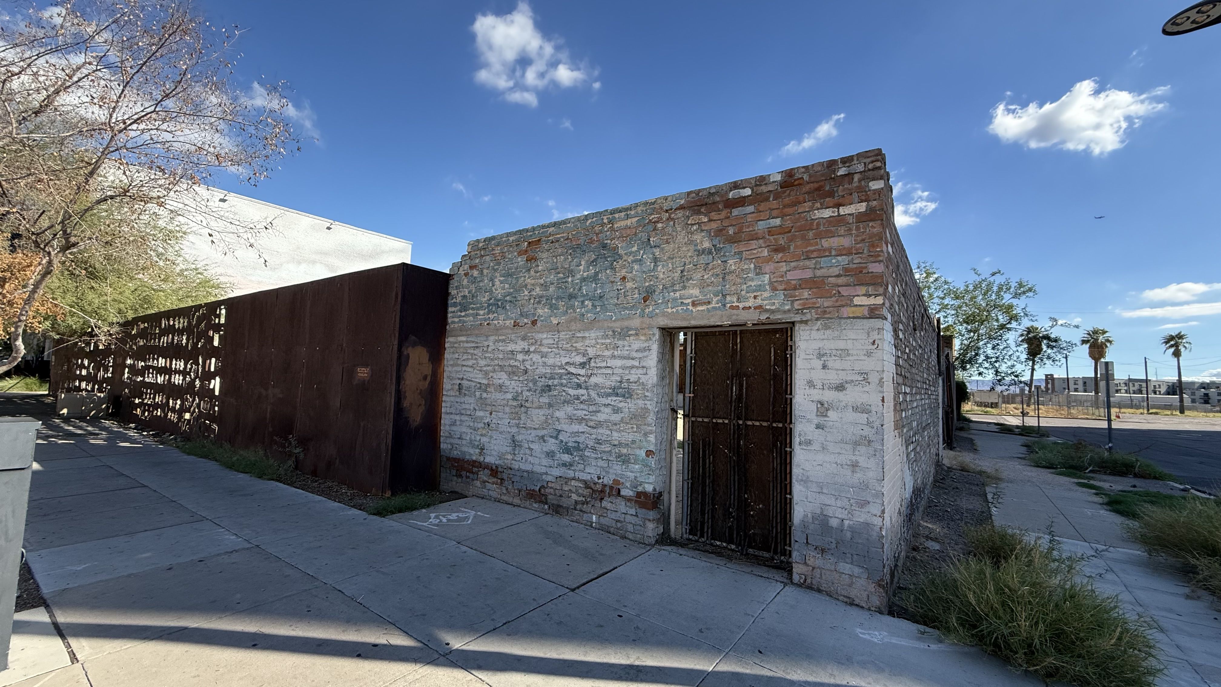 Wide street view of a small brick building with a whitewashed base and a rusted metal door, gate, along a concrete sidewalk; clear blue sky with a few clouds, bare tree on left, distant palm trees.