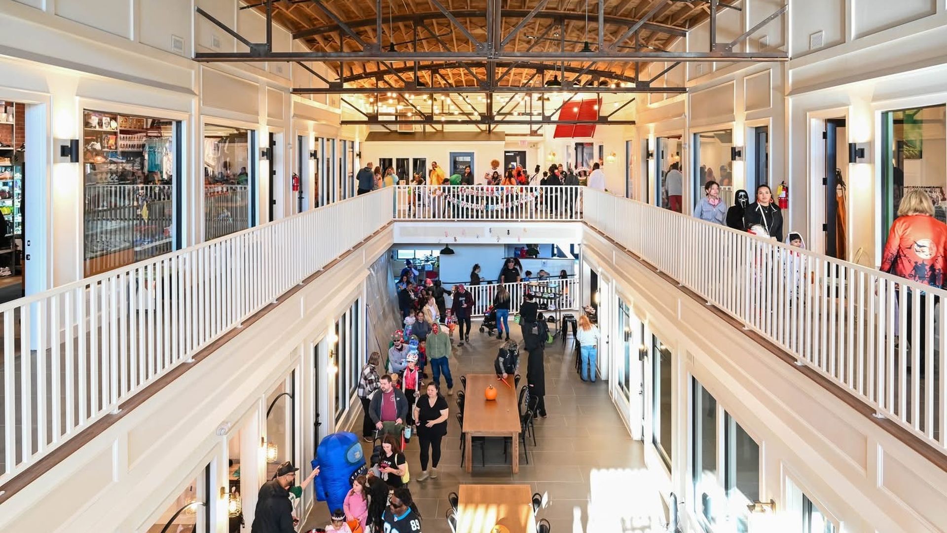 Two-story indoor space with white railings and wooden ceiling filled with people, some in costumes, walking along corridors and around rectangular tables with pumpkins.