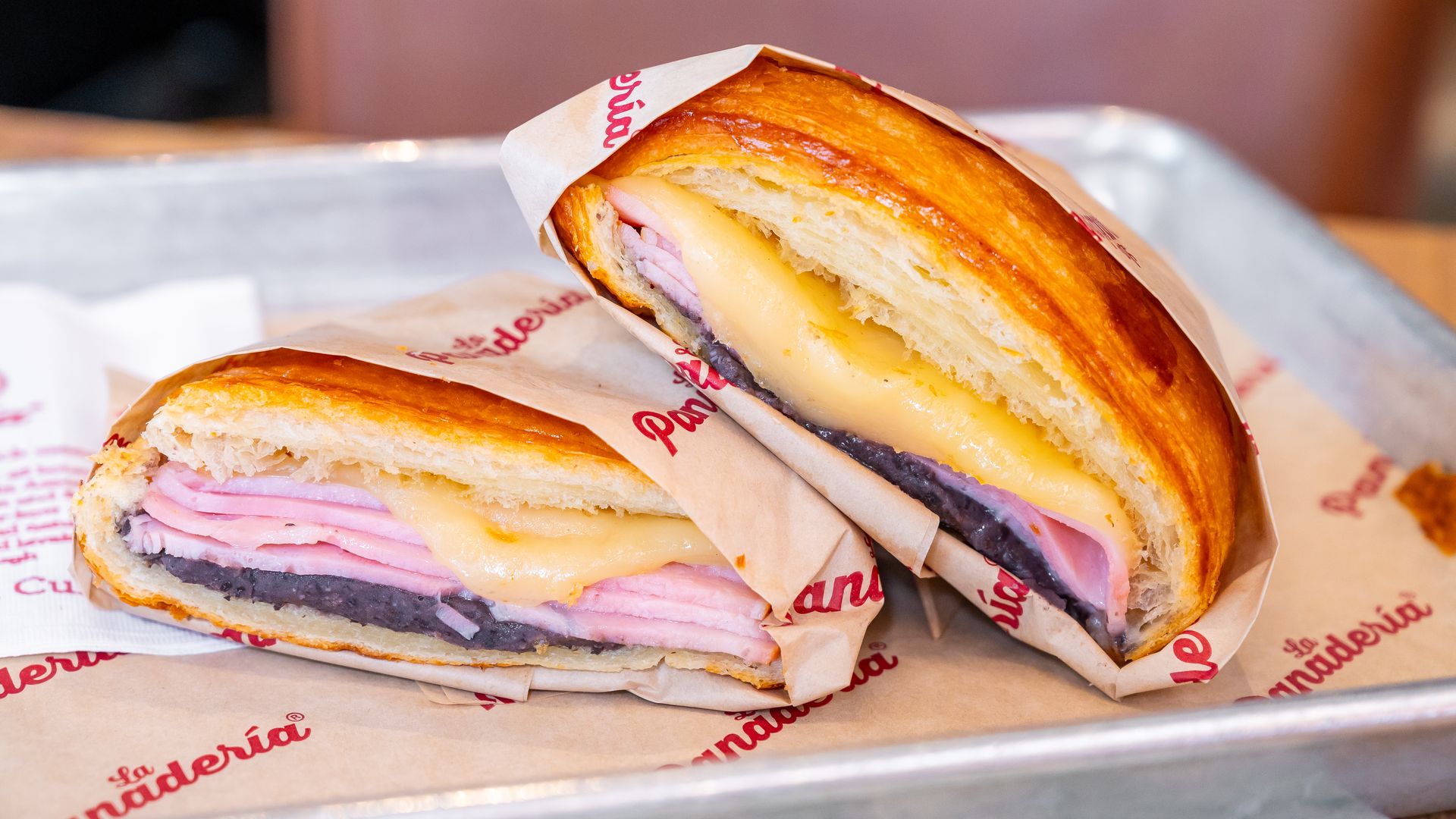 Close-up of a golden-brown croissant sandwich filled with pink ham and melted yellow cheese, wrapped in branded paper on a metal tray.