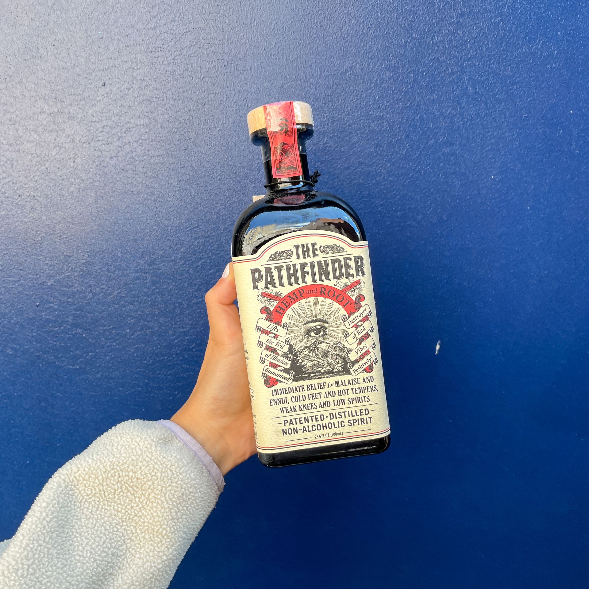 A hand holding up a bottle of The Pathfinder: Hemp & Root in front of a blue background.