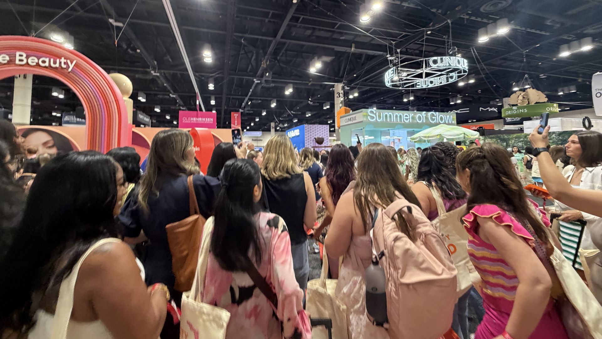 Back view of a crowded beauty expo, with attendees facing away, tote bags and backpacks. A pink circular arch on the left and a "Summer of Glow" booth sign overhead under bright ceiling lights.