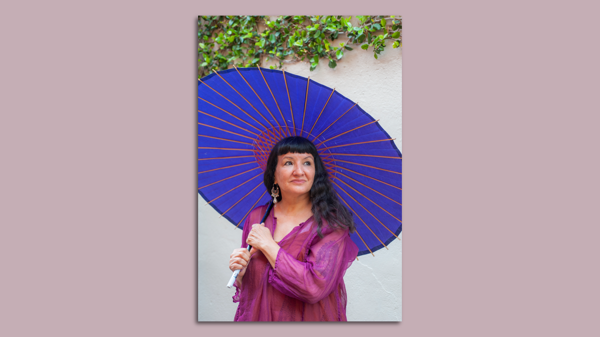 Sandra Cisneros is shown wearing a bright magenta top and holding a bright purple umbrella, against a white wall with green plants hanging above.