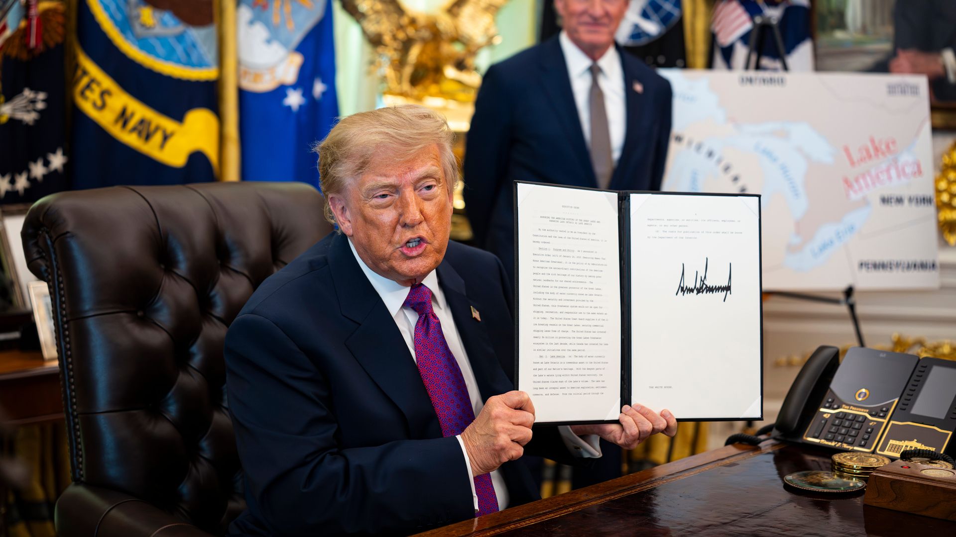 A man in a navy suit and red tie sits at a large wooden desk, presenting a two-panel document. Behind him are colorful flags, a gold eagle statue, and a map in a formal office setting.