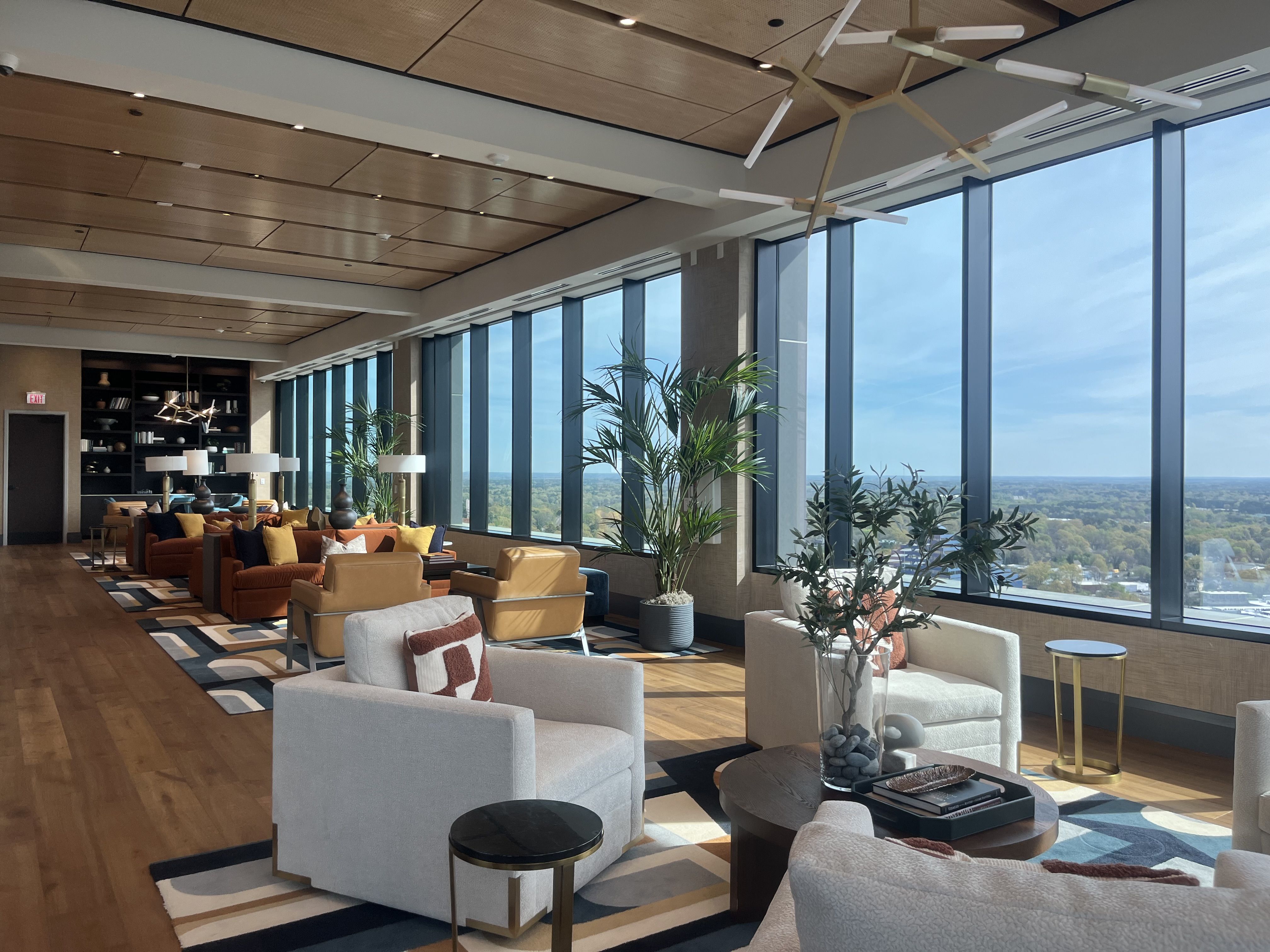 Spacious contemporary lounge with floor-to-ceiling windows offering a panoramic landscape and blue sky. Cream and tan seating, orange-brown sofas, colorful cushions, round coffee tables, potted plants, and a back bookshelf.