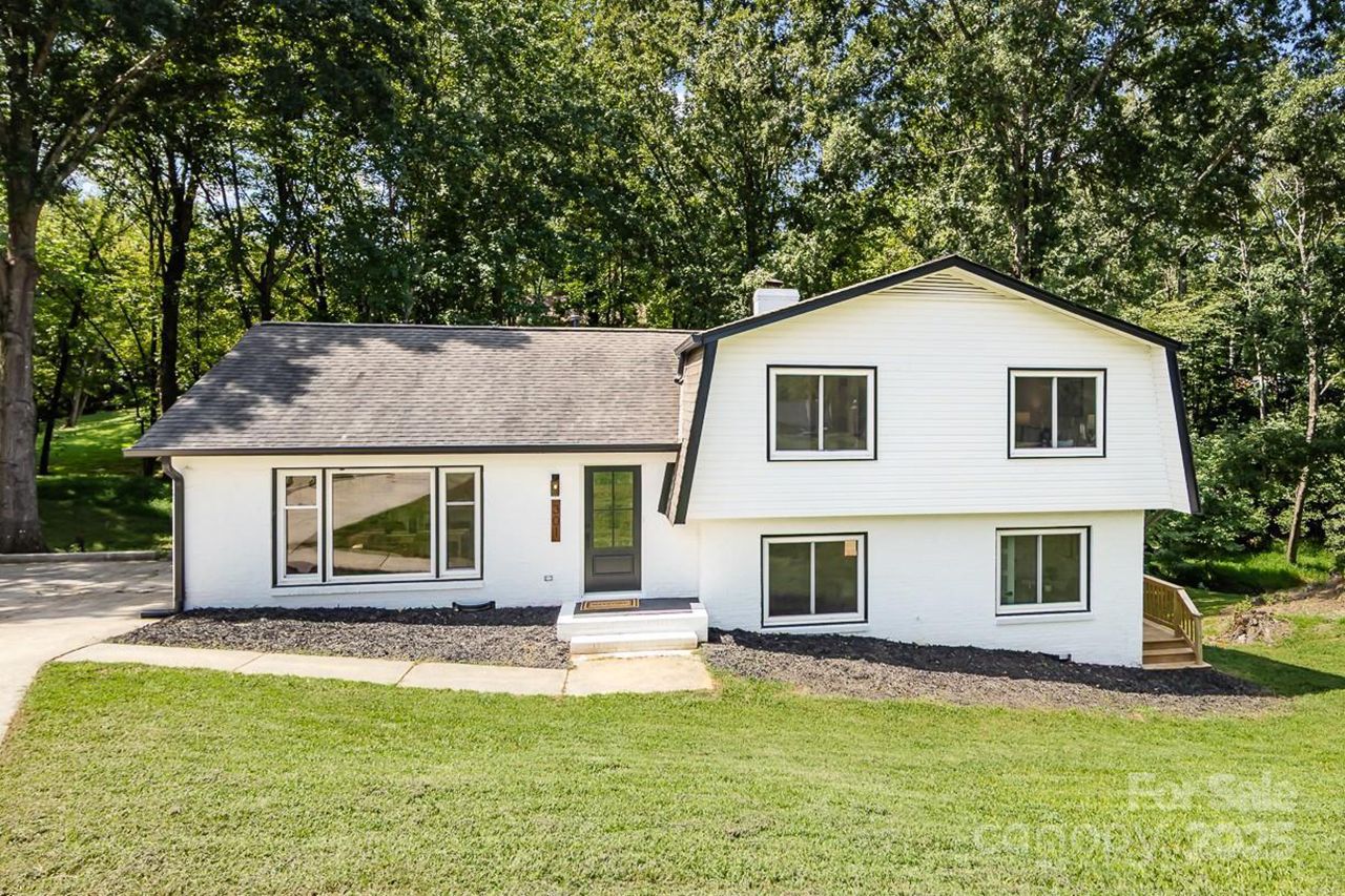 White two-story house with black-trimmed windows and gray front door, surrounded by green grass and tall trees in the background under a sunny sky.