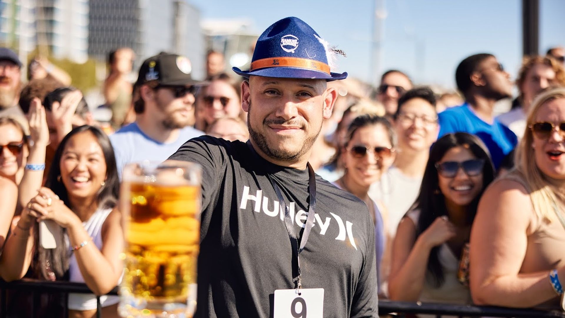 Smiling man wearing a blue hat and black Hurley shirt holds up a glass of beer at an outdoor event with a cheering crowd behind a barrier on a sunny day.