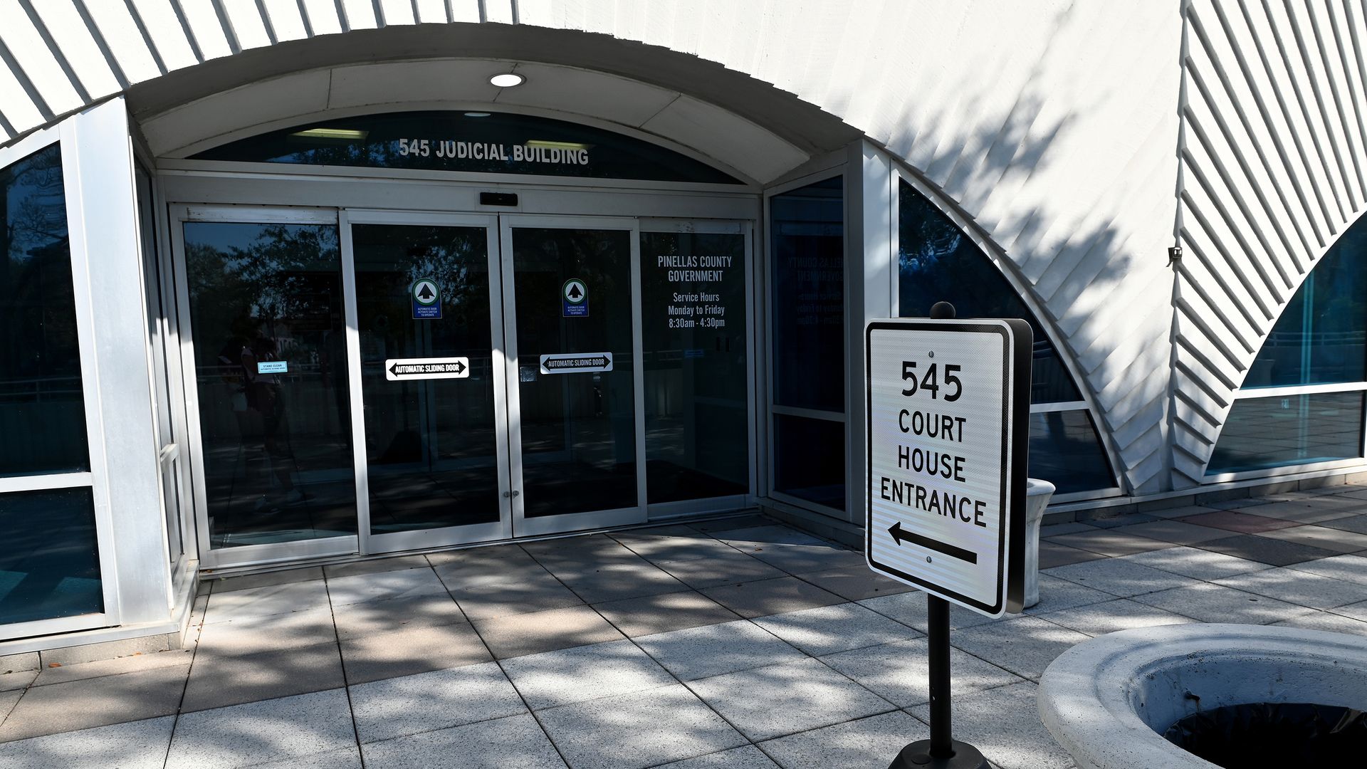 Entrance to a government building with glass automatic doors under a curved white facade; signs read "545 Court House Entrance" and "545 Judicial Building," with tree reflections in the glass.