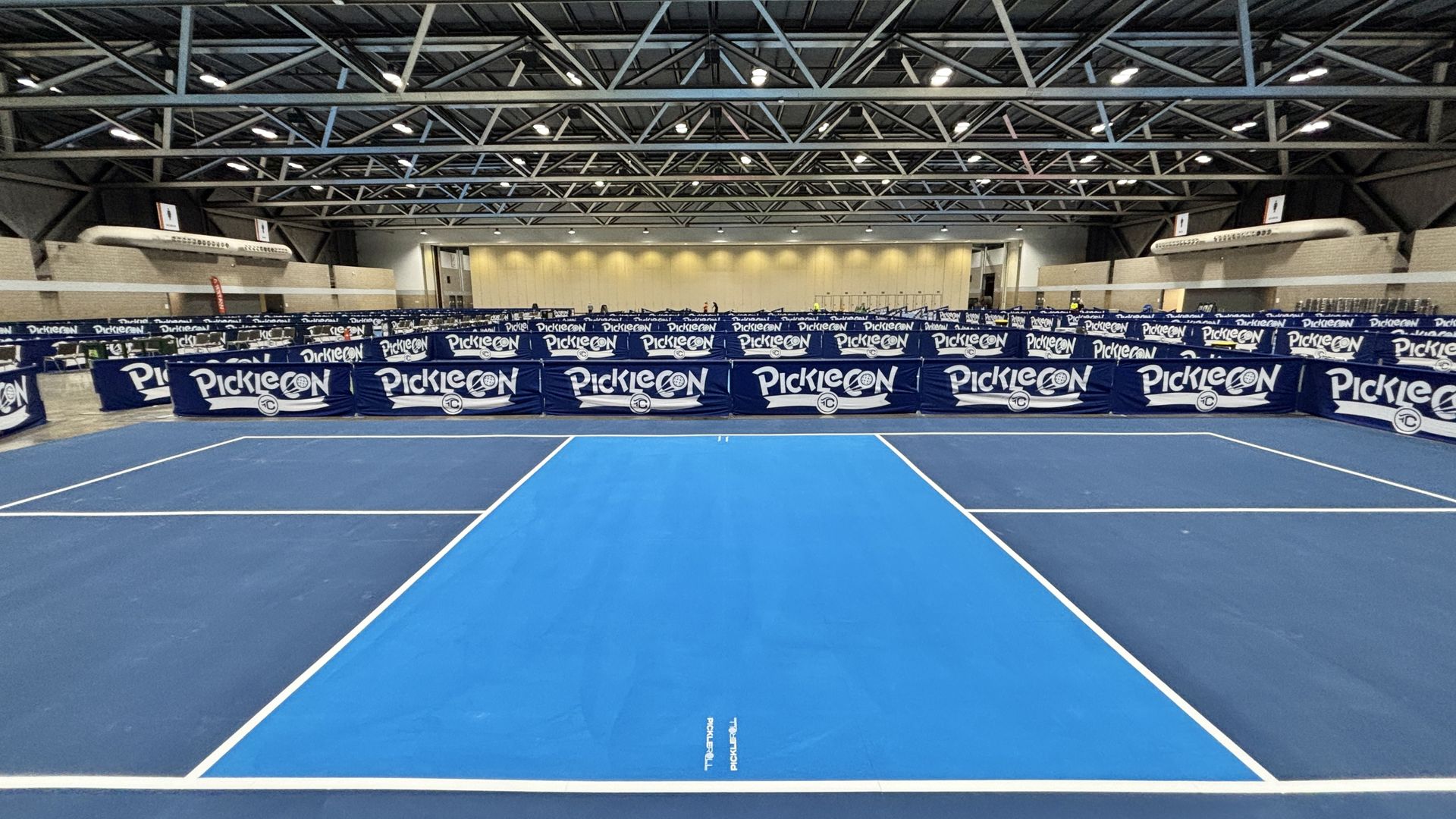 Indoor sports venue with rows of blue PickleCON barriers surrounding a pickleball court area. Bright overhead lights, exposed metal trusses, and a beige back wall in the far end.
