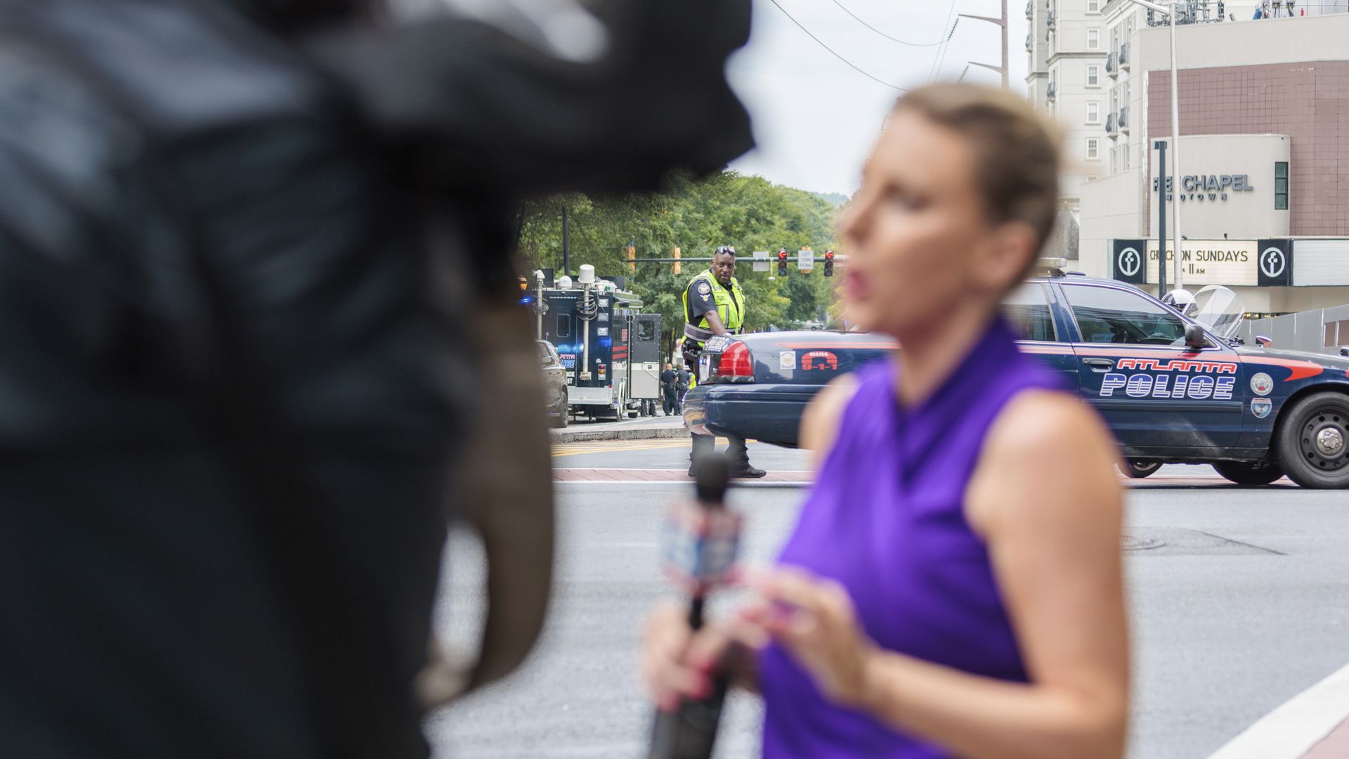 An out-of-focus TV journalist speaks to a camera as a police officer leans against a cruiser in an urban area in the background.